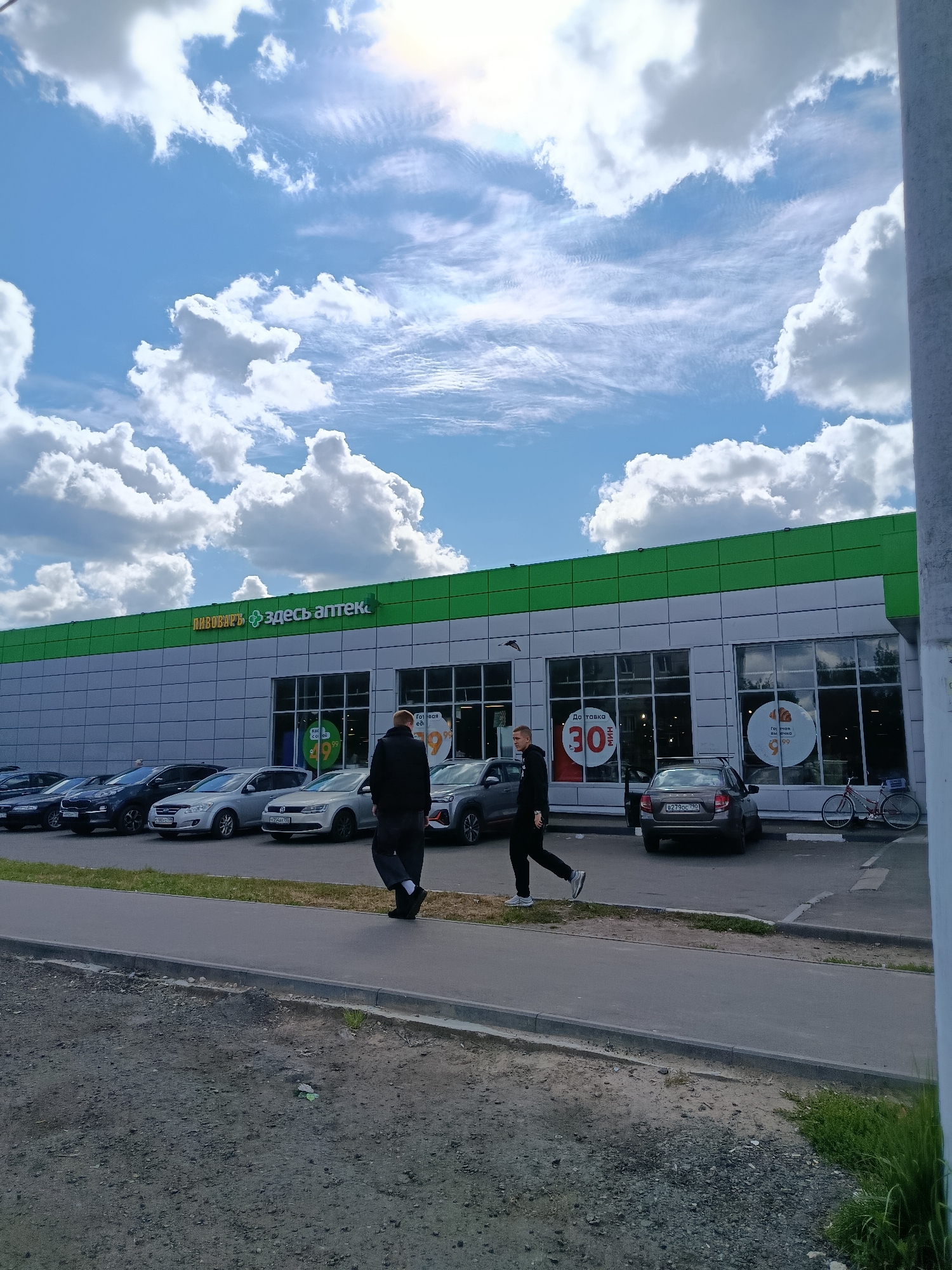 The image shows a street scene in the city of Orekhovo-Zuyevo, Russia, in front of a supermarket or pharmacy building. The building has a green and silver facade with large windows. A sign above the entrance reads "Пивоваръ Здесь аптека" (Pivovar Here pharmacy). Several cars are parked in front of the building. Two men are walking on the sidewalk. One man in a black jacket and pants is walking away from the camera. The other man, in a black tracksuit, is walking towards the camera. The sky is blue with white, fluffy clouds.