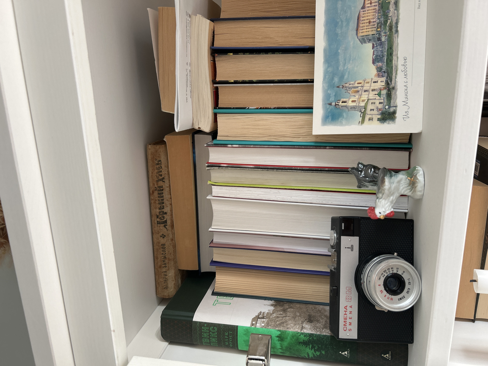 This image shows a shelf filled with books and a vintage camera. The books are stacked vertically and horizontally, with some spines visible. One book on the left has Russian text on its spine, appearing to be a title by "ВЛ. ЗАМЫСЛОВ". Another book in the foreground has green binding with Russian text that appears to be "ТВИН-ПИКC" and "МАРК ФОСТ". On top of the books, a postcard is placed, featuring a watercolor painting of buildings and a river, with Russian text "Из Минска с любовью" (From Minsk with love). A small ceramic rooster figurine is perched on a stack of books. A black film camera, identified as "СМЕНА 8М SMENA 8M" by visible text on its body, is positioned in front of some books. The overall setting appears to be an indoor shelf or bookcase. There are no people visible in the image. The lighting suggests an indoor environment, possibly daytime.