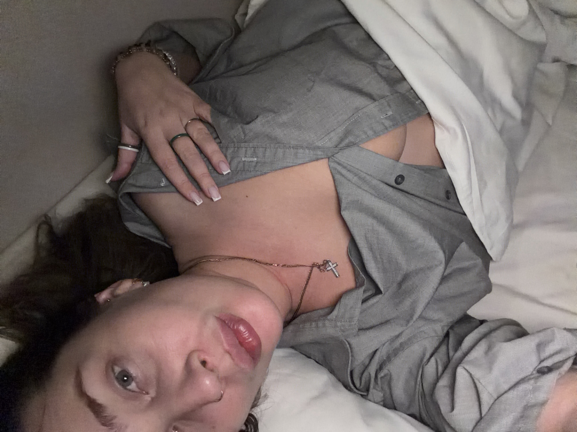 A person is lying on their back in a bed covered with white sheets and a gray button-down shirt. The person's head is turned slightly towards the viewer, revealing a partial view of their face. They have gray eyes, dark hair, and are wearing jewelry, including rings and a necklace with a cross pendant. The shirt is partially unbuttoned, revealing some cleavage. The background appears to be a simple indoor setting, likely a bedroom. There is no indication of a specific location in Moscow, Russia, or any other geographic context.