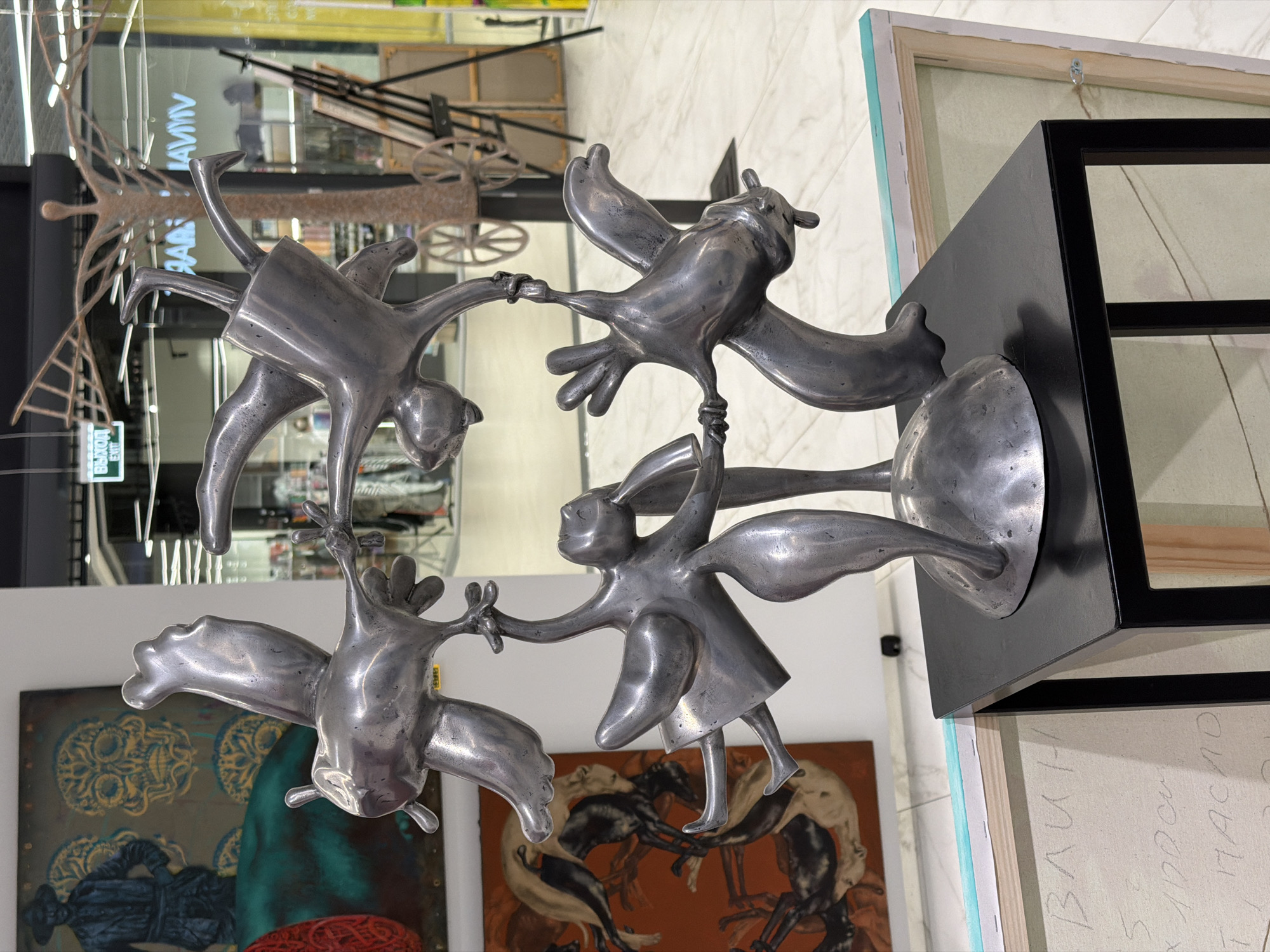 A metallic sculpture of four figures, resembling stylized birds or humanoid creatures, is displayed on a black pedestal in what appears to be an indoor public space, possibly an art gallery or a shopping mall in Minsk, Belarus. The sculpture is the central focus, with figures in dynamic poses, some inverted, appearing to be in motion or interacting with each other. Behind the sculpture, various artworks are visible: to the left, a large painting features a dark-clad figure and skull motifs; to the right, easels hold canvases, one with a textured, tree-like structure. Further in the background, a store with a bright yellow facade and a sign reading "VIVARIUM" is visible, along with a large window reflecting an interior space with shelves and lighting. An "EXIT" sign is also present above a doorway. The floor is a polished, light-colored tile. The lighting suggests an artificial indoor environment.