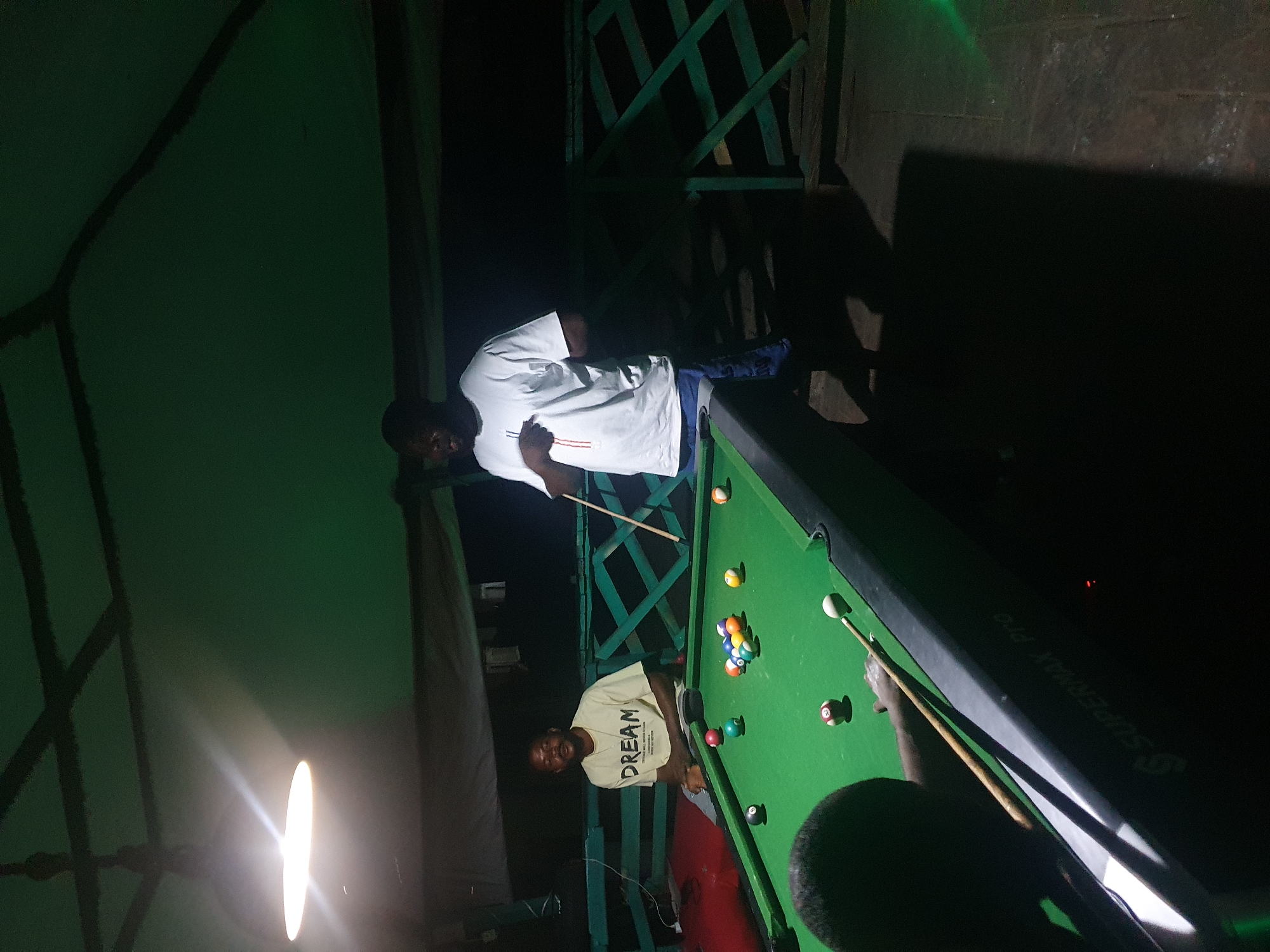 Two men are playing pool in an outdoor, covered area. The man on the left is sitting down and wearing a light-colored t-shirt with "DREAM" printed on it. The man on the right is standing and wearing a white t-shirt and blue pants, holding a pool cue. A pool table with several balls is in the foreground. The environment appears to be a casual, recreational space, possibly at a bar or private residence, located in Lokoja, Nigeria, suggested by the style of the structure and the ethnicity of the individuals.