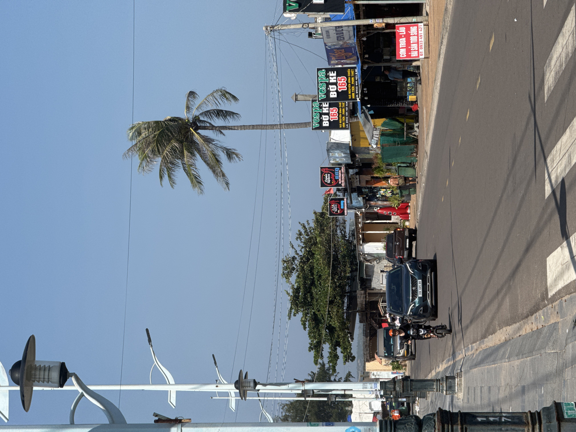 The image depicts a street scene in Mũi Né, Vietnam, characterized by a bright, sunny day with a clear blue sky. A paved road with visible white pedestrian crossings runs through the center of the frame, with traffic moving along it.
On the left side of the road, a sidewalk is bordered by decorative lamp posts and a low wall. Further down the sidewalk, a person on a small motorcycle, wearing a dark shirt and a helmet, is seen riding towards the viewer. Behind the motorcycle, a dark-colored SUV is following.
The right side of the road features a row of commercial establishments, including shops and restaurants, identifiable by various signs. Notably, a prominent palm tree with fronds blowing in the wind stands tall against the sky, suggesting a coastal or tropical environment. Several signs with Vietnamese text are visible, including ones advertising "Vespa Bò Kê" and "Cơm Trưa - Lẩu Hải Sản Tươi Sống". Some people are seen walking near these establishments. The overall impression is of a lively street with ongoing commercial activity under a clear sky.