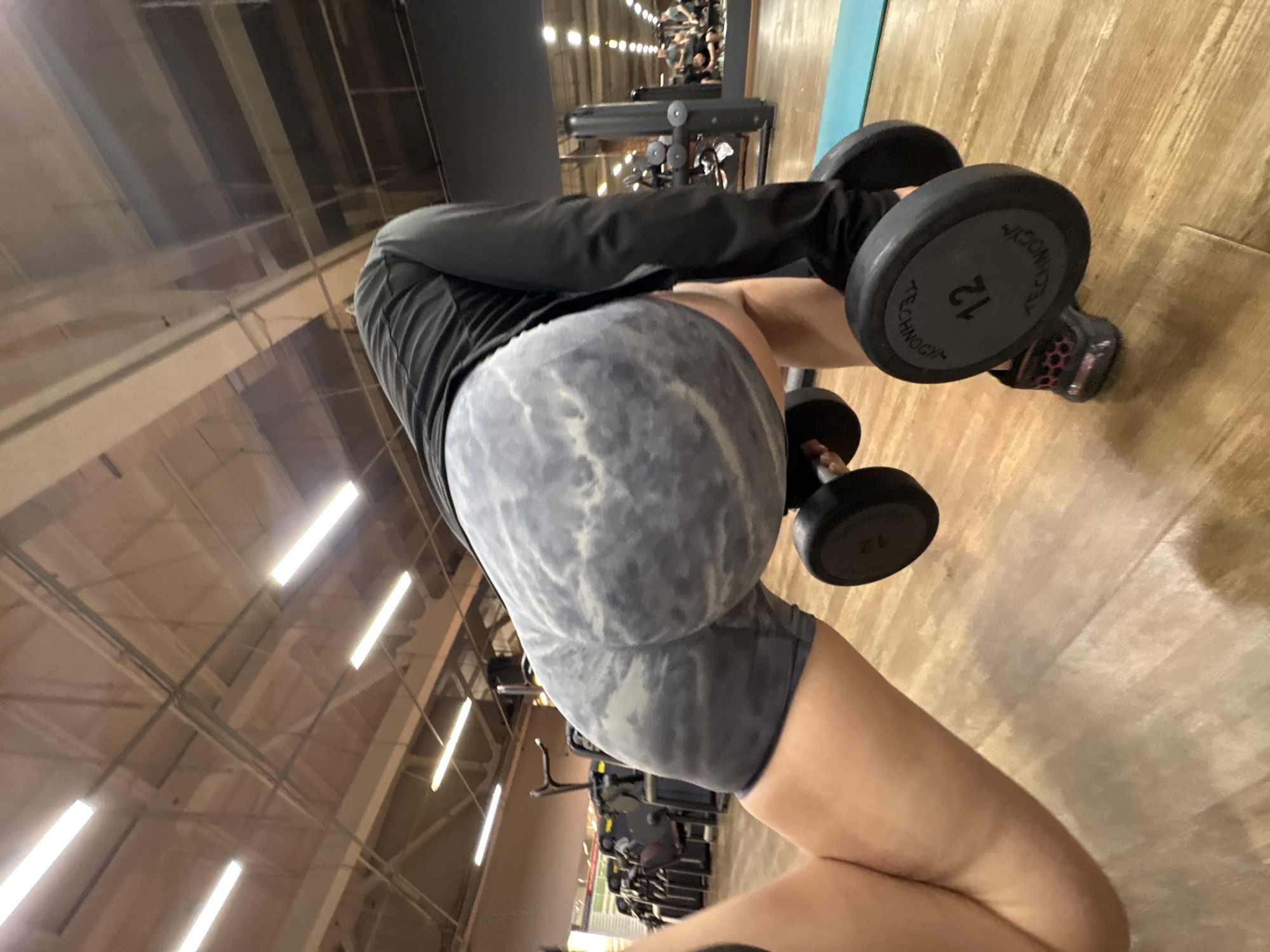 The image depicts a person in a gym setting, holding dumbbells. The person is wearing dark, long-sleeved athletic wear and gray and white patterned shorts. The dumbbells are black, one with the number "12" visible on the side. The flooring is light brown wood. In the background are other gym machines and people. The location is likely a gym in Chaikovsky, Russia.