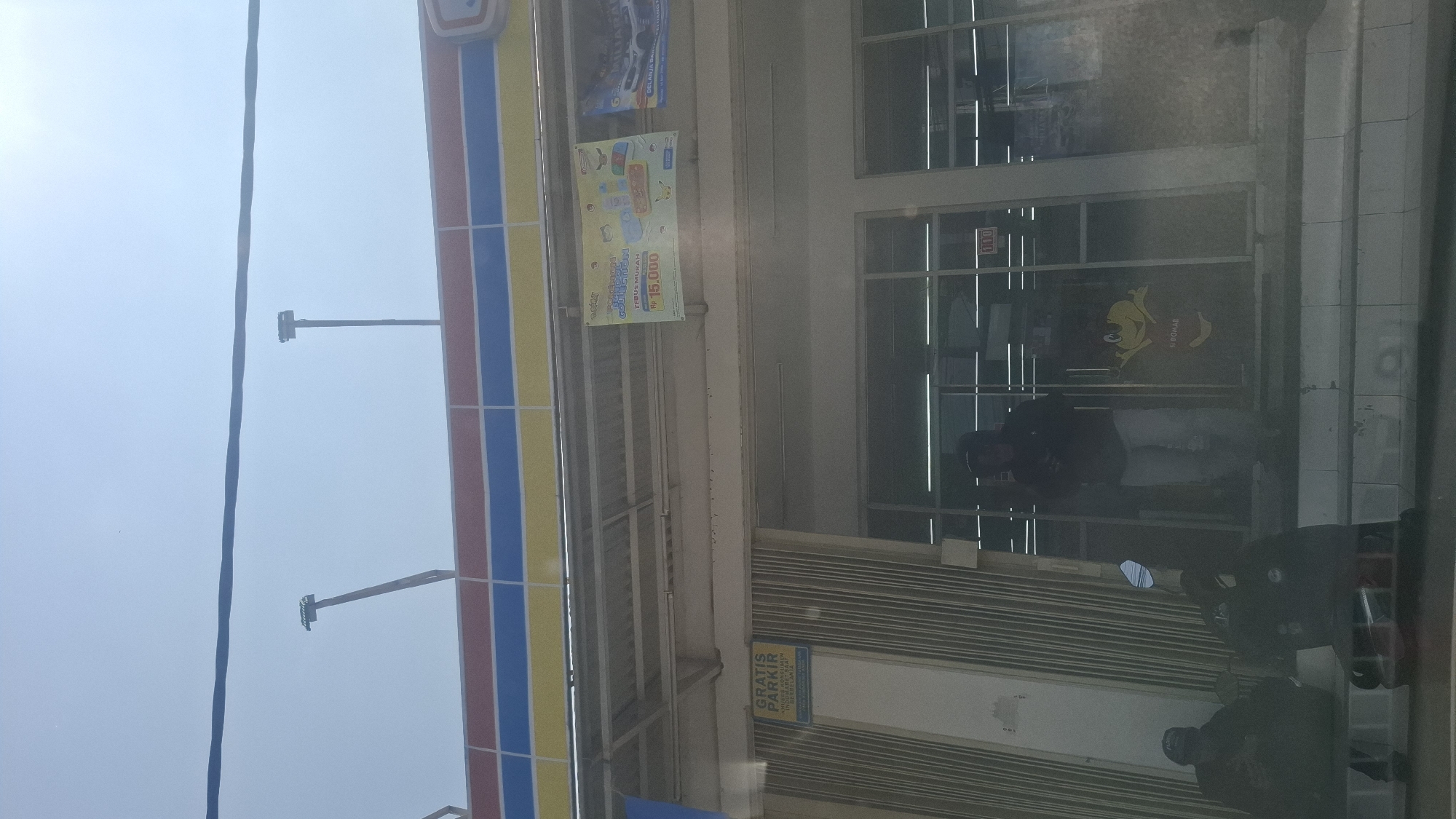 The image shows a storefront in Rancagoong, Indonesia, under a bright, clear sky. The building has a colorful facade with horizontal stripes of red, yellow, and blue. Above the building, two tall, thin poles extend upwards, one leaning slightly to the left. A dark power line runs horizontally across the top of the image.
In front of the storefront, there are two distinct sections. To the left, a vertical sign reads "GRATIS PARKIR," meaning "Free Parking," with additional text below. Next to this sign is a ribbed, metallic shuttered door.
The main entrance to the storefront is a set of glass doors, through which several individuals are visible. One person, wearing a dark top and light-colored pants, stands inside the doorway. Another person, wearing a dark hooded jacket and a cap, is seated outside the entrance to the left, accompanied by another individual similarly dressed. A motorcycle is parked to the left of the entrance.
A poster is displayed on the building to the right of the shuttered door, featuring cartoon characters and text that appears to be an advertisement. A yellow cartoon character is visible on the glass door. The overall scene depicts a commercial area on a sunny day.