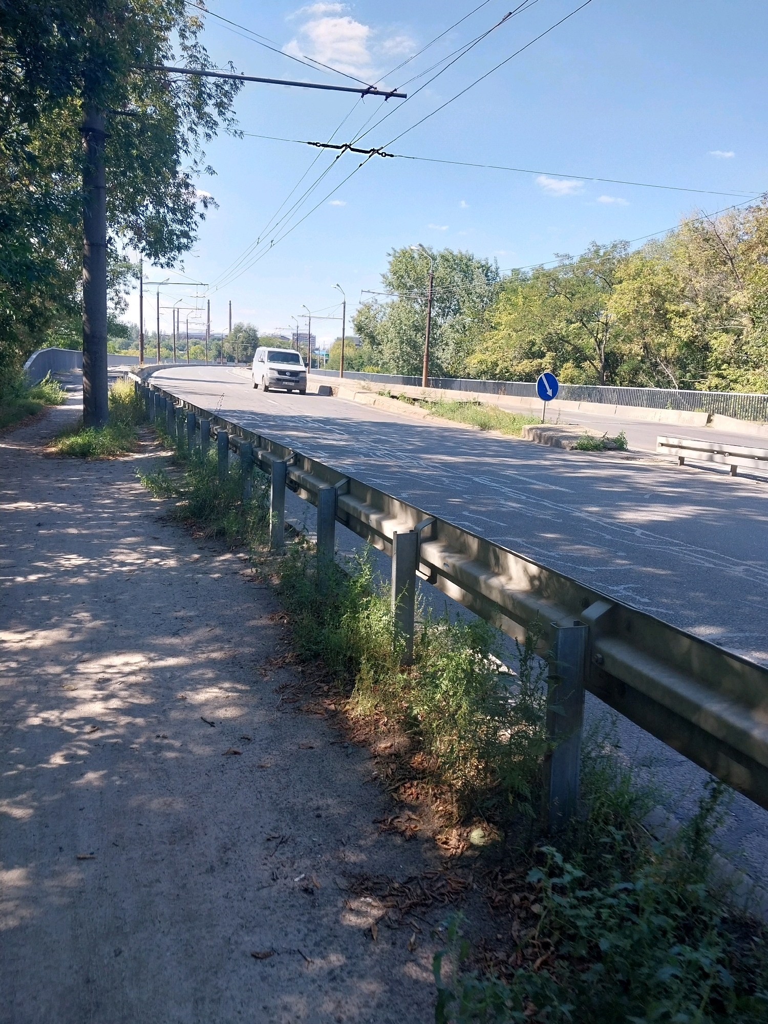 The image depicts a street scene in Dnipro, Ukraine, featuring a road with a white van driving on it. Overhead, trolleybus wires are visible, indicating the presence of public transportation infrastructure. The road is bordered by a metal guardrail and a sidewalk, with vegetation growing along the roadside. Trees line the background, and the sky is partly cloudy. A blue traffic sign with a white arrow pointing downwards is also visible on the side of the road. The scene appears to be captured during daytime, with sunlight casting shadows on the sidewalk.