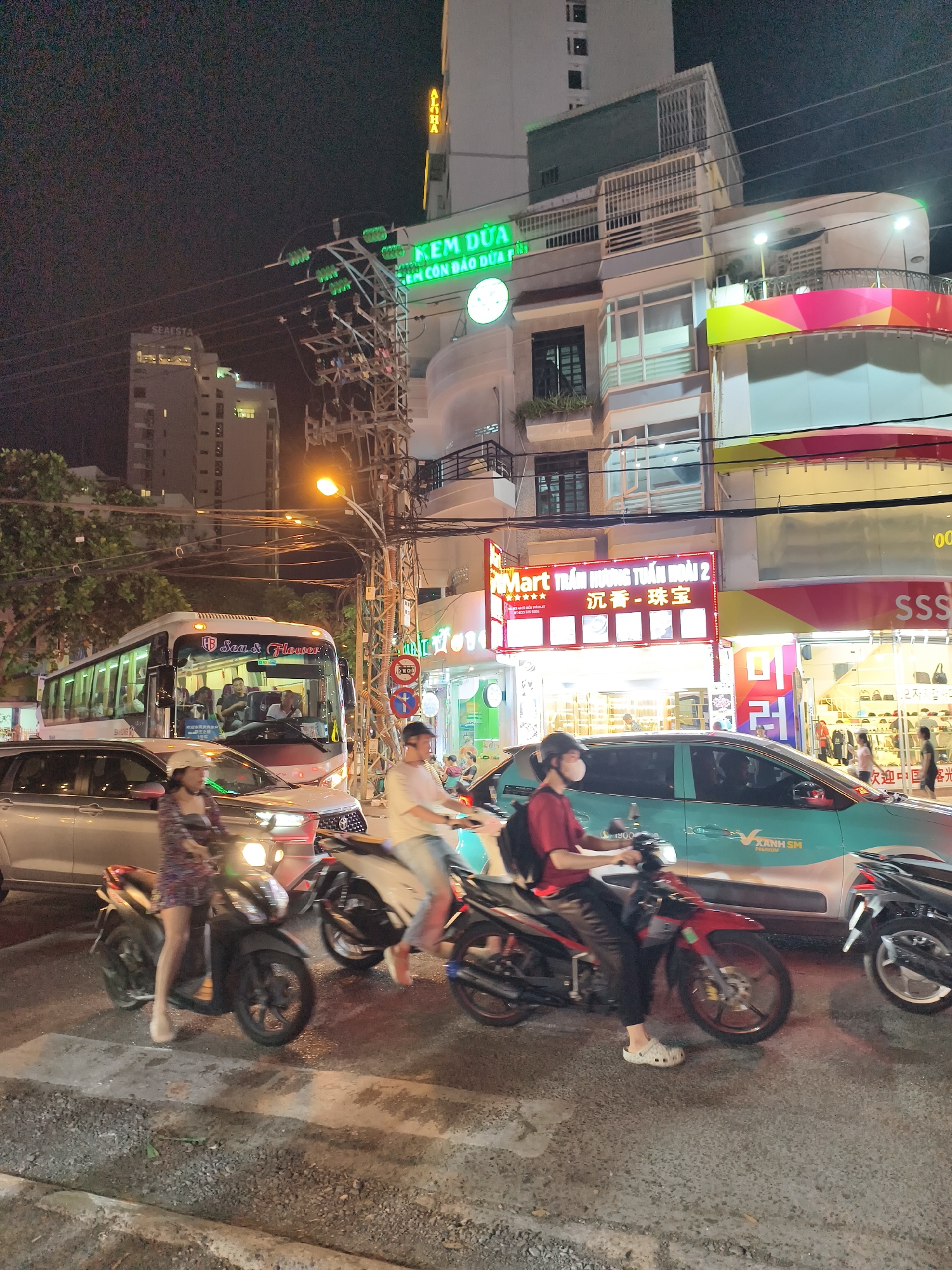 This image depicts a nighttime street scene in Nha Trang Ward, Vietnam, characterized by artificial illumination from streetlights, vehicle lights, and numerous commercial signs.
In the upper-left, a tall building bears the illuminated name "SeAESTA." To its right, another building features a prominent green sign with Vietnamese text "KEM DUA" (Coconut Ice Cream) and a vertically oriented yellow sign displaying "TOHA." Further right, a building exhibits a vibrant red and yellow facade, below which a brightly lit red sign reads "KMART TRẦM HƯƠNG TUẤN HOÀN 2," with a partial "SSS" sign visible nearby. A dense network of overhead power lines and a utility pole equipped with an orange-glowing streetlight occupy the center of the frame.
Traffic is active on the asphalt road surface. A white and red bus, marked "HB Sea & Flower," is visible in the midground, with multiple passengers seated inside. Directly in front of the bus is a silver SUV with a visible front grille. To the right of the bus, a teal-colored sedan, identified as an electric taxi by "XANH SM PREMIUM" and "1900" branding on its side, is positioned.
Multiple individuals are present, primarily on motorbikes. In the lower-left, a female wearing a patterned dress and light-colored footwear is riding a dark-colored scooter. Central to the foreground, a male in a light-colored shirt and pants operates a white scooter. To the right, a male in a red shirt, black pants, a face mask, and white Crocs, carrying a backpack, is riding a red motorbike. Other motorbikes are partially visible throughout the foreground. Pedestrians are discernible on the sidewalk adjacent to the commercial establishments in the midground. Traffic signs, including one with "2 CHIỀU" (Two-Way) text, are affixed to a pole to the right of the bus.