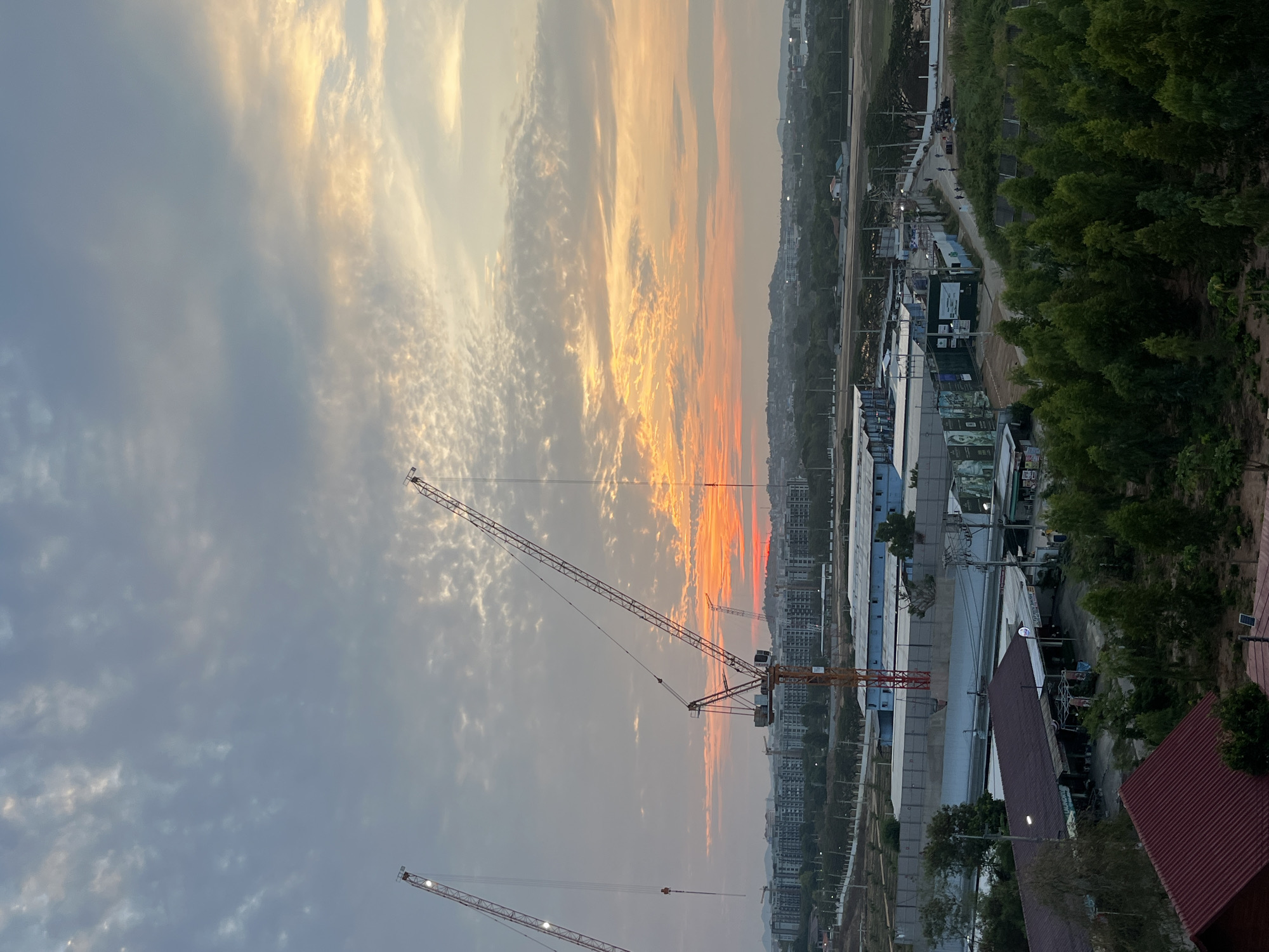 The image depicts a cityscape in Pattaya City, Thailand, during a sunset. The sky is filled with clouds illuminated by orange and golden light. Several construction cranes are visible, indicating ongoing development in the area. Buildings and infrastructure line the lower part of the image, transitioning into a green, forested area in the upper right. A road with occasional vehicles is also discernible.