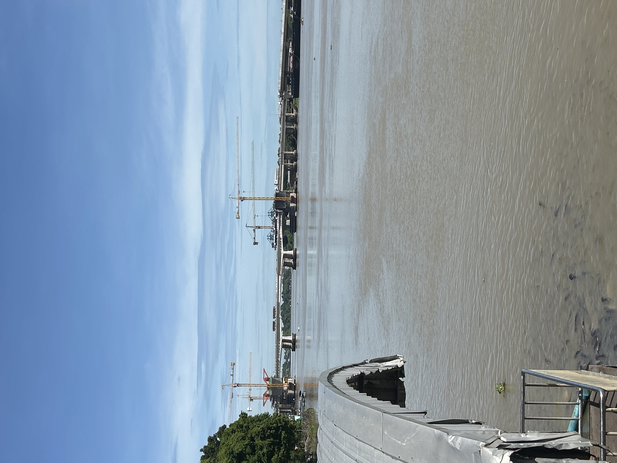A wide, muddy river flows under a bright blue sky with scattered clouds. In the foreground, the side of a grey, curved structure is visible, extending from the left edge of the frame. A metal railing and a blue pipe are attached to it. Below the railing, a large school of dark fish swims in the murky water.
Across the river, a large bridge construction project is underway. Several yellow cranes stand tall, with two prominent ones near the left bank. The bridge spans the width of the river, with concrete pillars supporting sections of the roadway. Some completed sections of the bridge are visible, and others appear to be under construction. A barge is moored to the right side of the bridge's completed section. Lush green trees line the distant bank of the river. The scene suggests a bustling construction site in a semi-urban or natural environment. The location is identified as Thai Ko Subdistrict, Thailand.