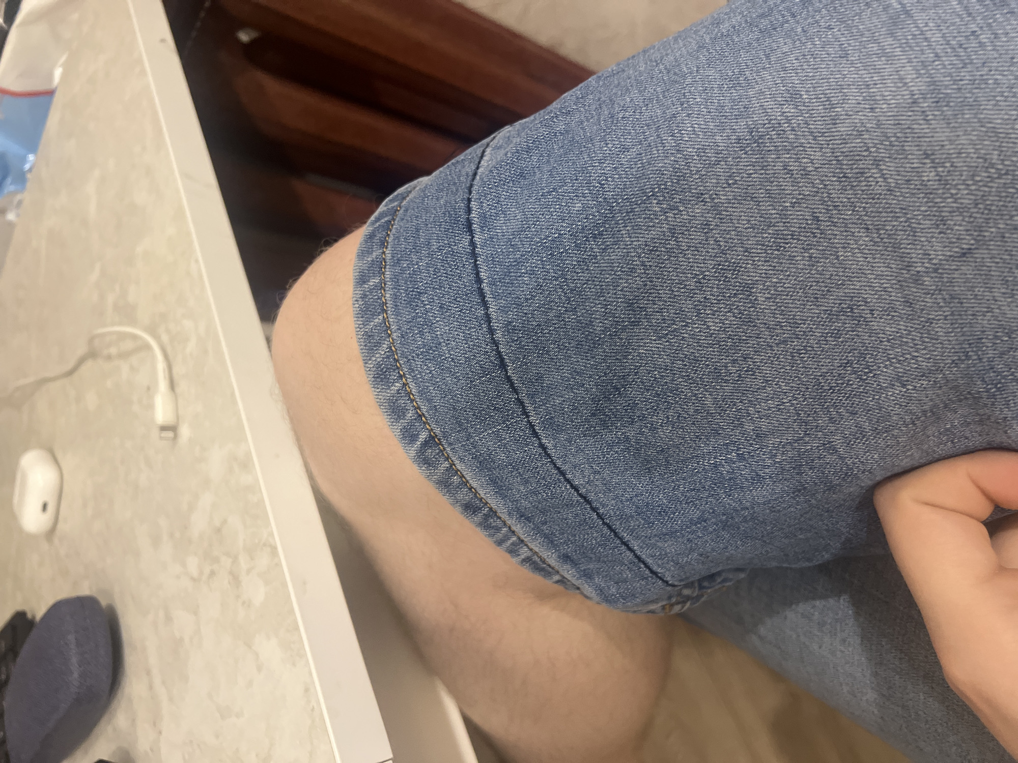 The image shows a close-up view of a person's legs, wearing denim shorts, and a hand resting on their leg. The setting appears to be indoors, possibly at a desk, with a portion of a countertop visible in the background. On the countertop, there are several objects including what appears to be a charging case for earbuds and a white charging cable. The person's legs are bare above the shorts, and the texture of the denim is clearly visible. The lighting suggests an indoor environment. There is no visible indication of the location being in Saratov, Russia.
