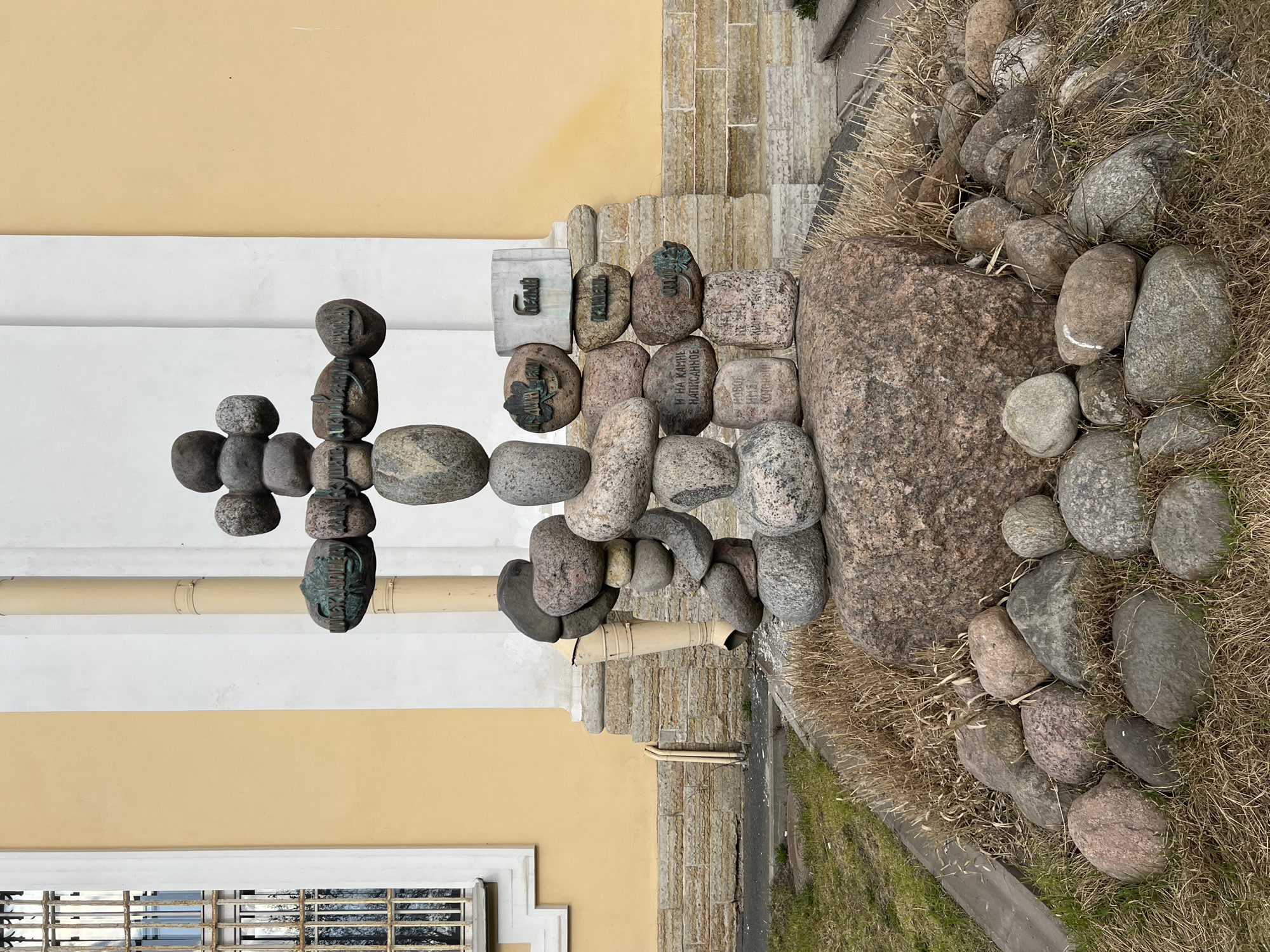 The image depicts a memorial cairn shaped like an Orthodox Christian cross, constructed from numerous rounded natural stones of varying sizes and shades of grey and pinkish-grey. This monument is located in Troitskaya Square (Trinity Square) in Saint Petersburg, Russia, opposite the Solovetsky Monastery Representation Church.
The cross rests on a large, irregular reddish-brown boulder, which is surrounded by smaller, similarly colored stones embedded in sparse, dry, yellowish-brown grass interspersed with some green shoots. Several stones incorporated into the cross structure feature dark metal plaques with Cyrillic inscriptions and embossed leaf motifs. Additional stones at the base of the cross bear carved Cyrillic text.
In the background, a two-toned building facade is visible: light yellow on the left and white on the right. A prominent cream-colored vertical drainpipe extends down the white section of the wall. To the far left, a window with white trim and rusted vertical metal bars is set into the yellow wall, reflecting bare tree branches. The building's foundation is composed of horizontally stacked, light-colored stone blocks. A concrete curb and a sliver of dark pavement are present in the lower left, with a partially obscured metal manhole cover visible in the grass. The scene is illuminated by diffused light, and no individuals are present.
