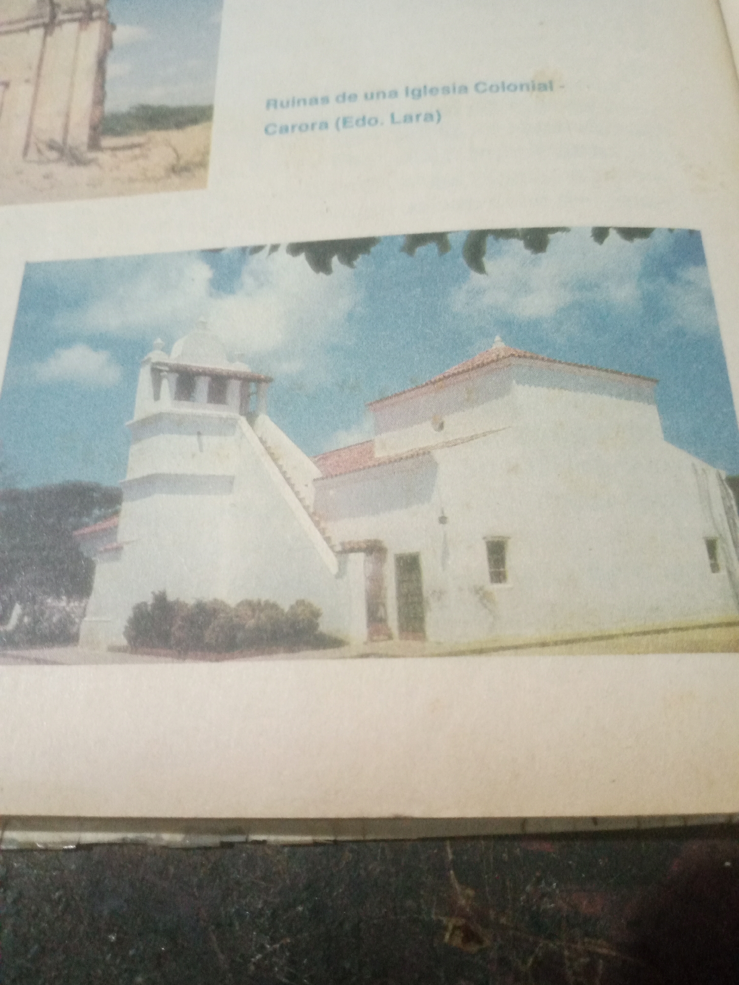 The image displays a historical building, likely a church, with white walls and a terracotta tiled roof. A prominent bell tower with arched openings stands to the left, adjacent to a staircase leading to an upper level. The building is situated in an outdoor environment with some greenery and trees in the foreground and background, under a cloudy blue sky. Text overlays on the image read "Ruinas de una Iglesia Colonial - Carora (Edo. Lara)". This suggests the building is the ruins of a colonial church located in Carora, Lara state, Venezuela.
