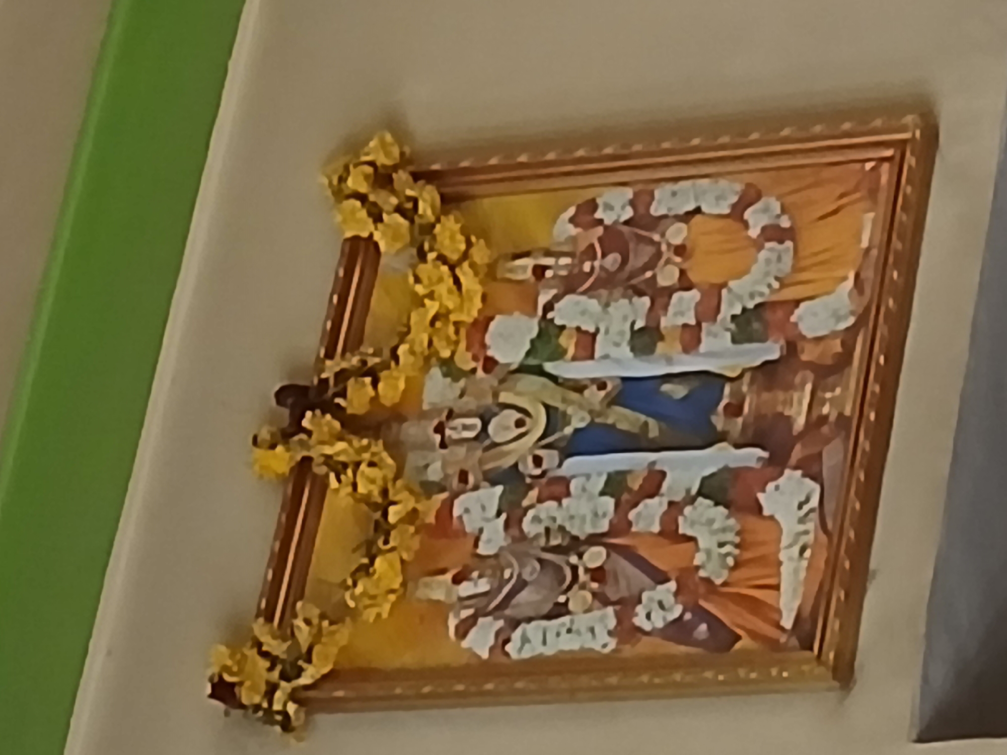 The image shows a framed painting of Hindu deities, specifically Lord Shiva and Goddess Parvati, adorned with garlands of yellow flowers. The painting is mounted on a wall, likely in a Hindu household. The background is a plain wall with a vertical green edge on the left, possibly a wall corner. The image lacks sufficient context to determine a specific location within Bengaluru, India.