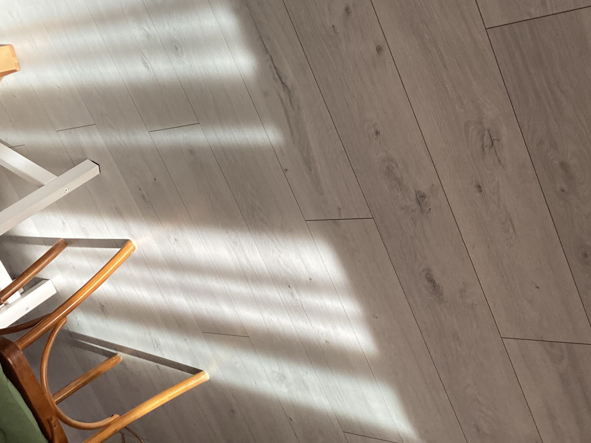 The image shows a close-up view of a light-colored wooden floor. Sunlight streams in from the left, creating diagonal stripes of light and shadow across the planks. The wooden planks are arranged diagonally, with visible grain patterns and knots. In the upper left corner, the legs of a wooden chair with a green seat are visible, casting a distinct shadow onto the floor. To the right of the chair legs, part of a white object, possibly a table leg, is also present. The overall impression is that of an indoor setting with natural light.
