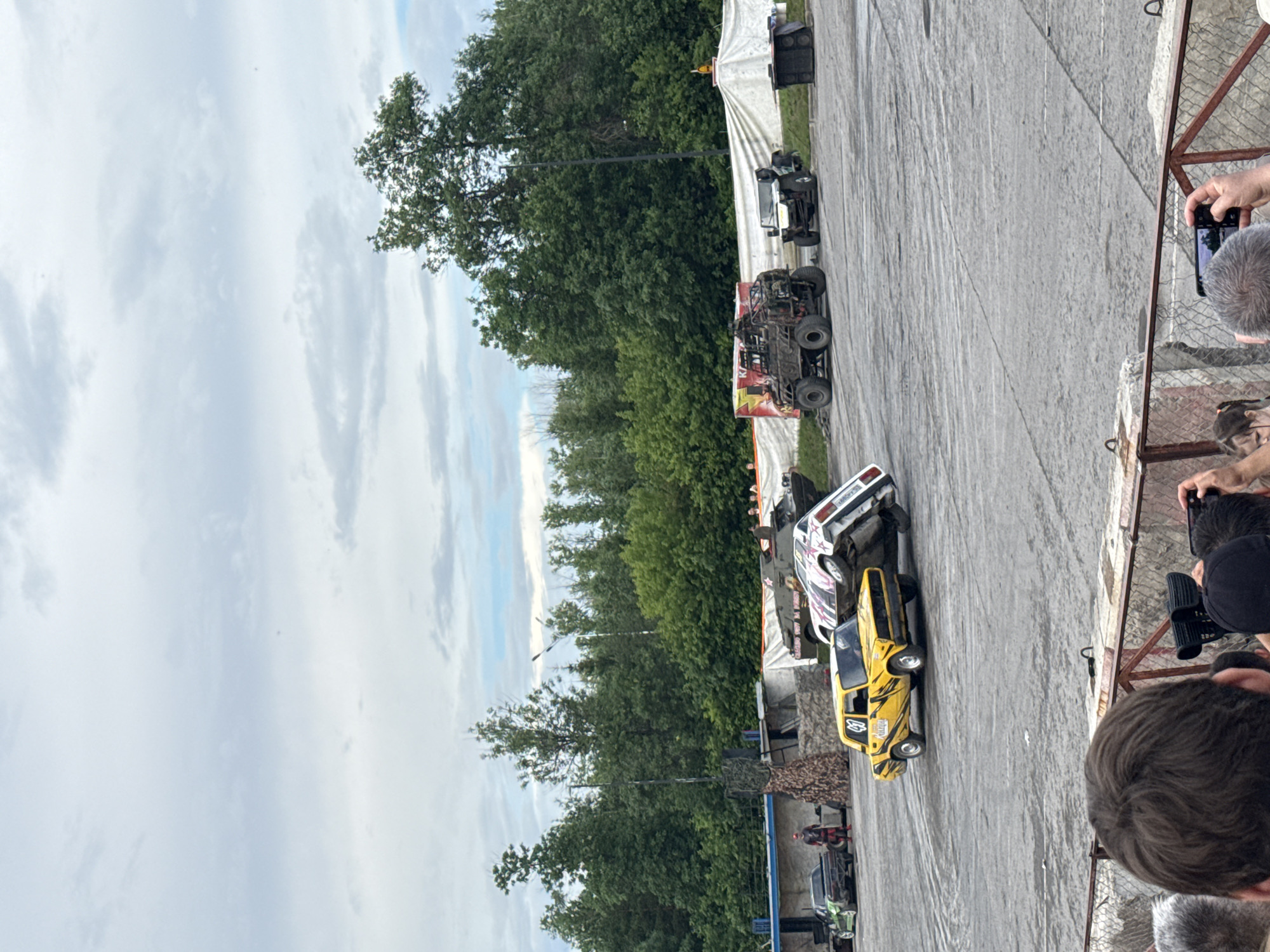 An outdoor stunt show is in progress in Unknown, Unknown. The scene features a variety of vehicles, including a yellow car, a white car, a large military-style vehicle, and a white jeep. The yellow car is in the foreground, appearing to be driving. Behind it, the white car is on its side, possibly mid-stunt. The military vehicle and jeep are parked in the background. Several trees and a cloudy sky form the backdrop. In the foreground, spectators are visible, some holding cameras and phones, indicating they are observing the event. A person in a red outfit is standing near a black car on the left side of the image. There are also various structures and equipment associated with a stunt show setup, such as a canvas barrier and what appears to be a sound system.