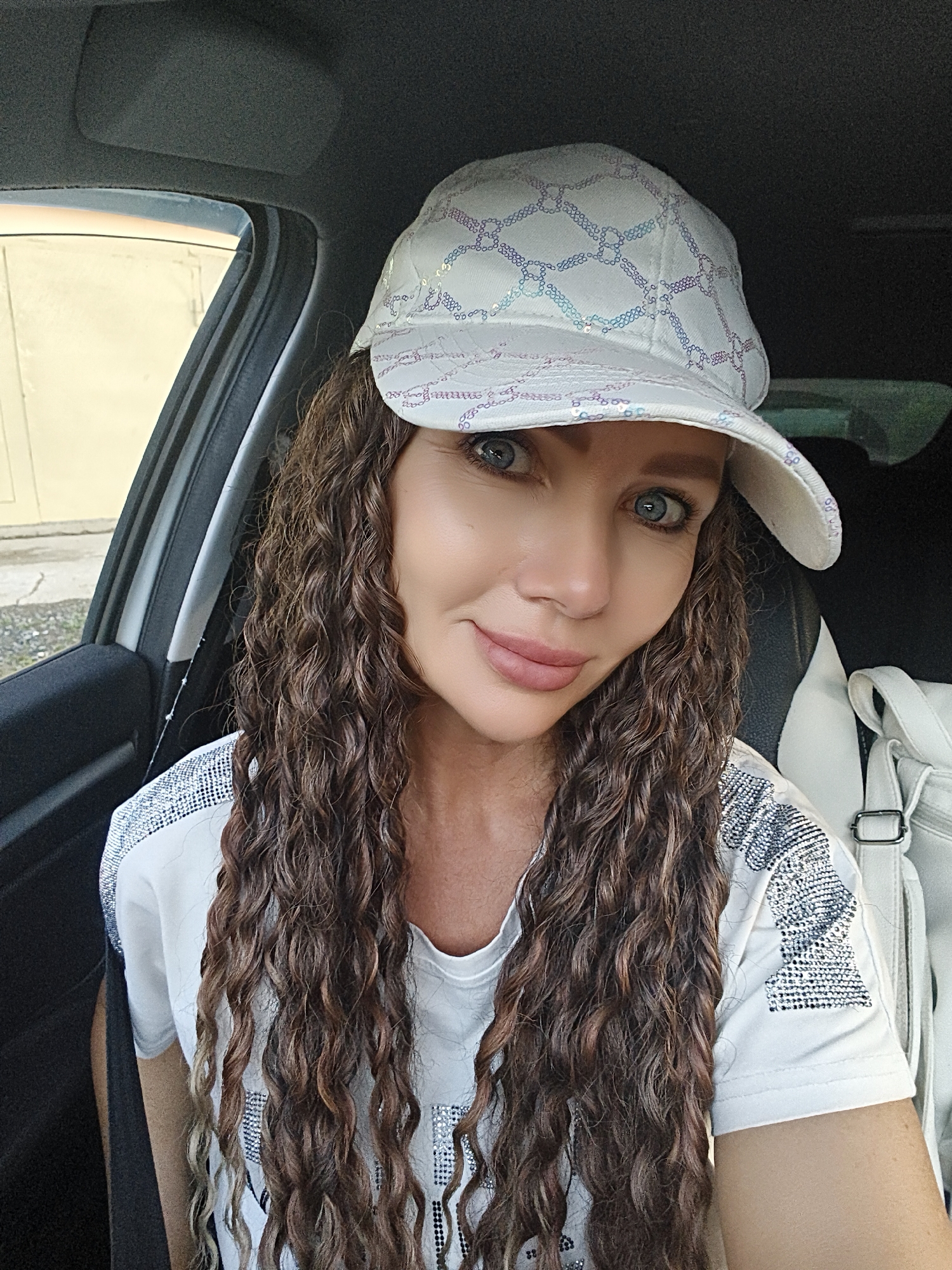 A woman with long, curly brown hair and blue eyes is sitting in the passenger seat of a car. She is wearing a white baseball cap with a diamond pattern and a white t-shirt with sparkling embellishments on the shoulders. She is smiling and looking at the camera. A white backpack is visible behind her. The background shows the interior of the car and a blurred view of the outside. The location is Unknown, Unknown.