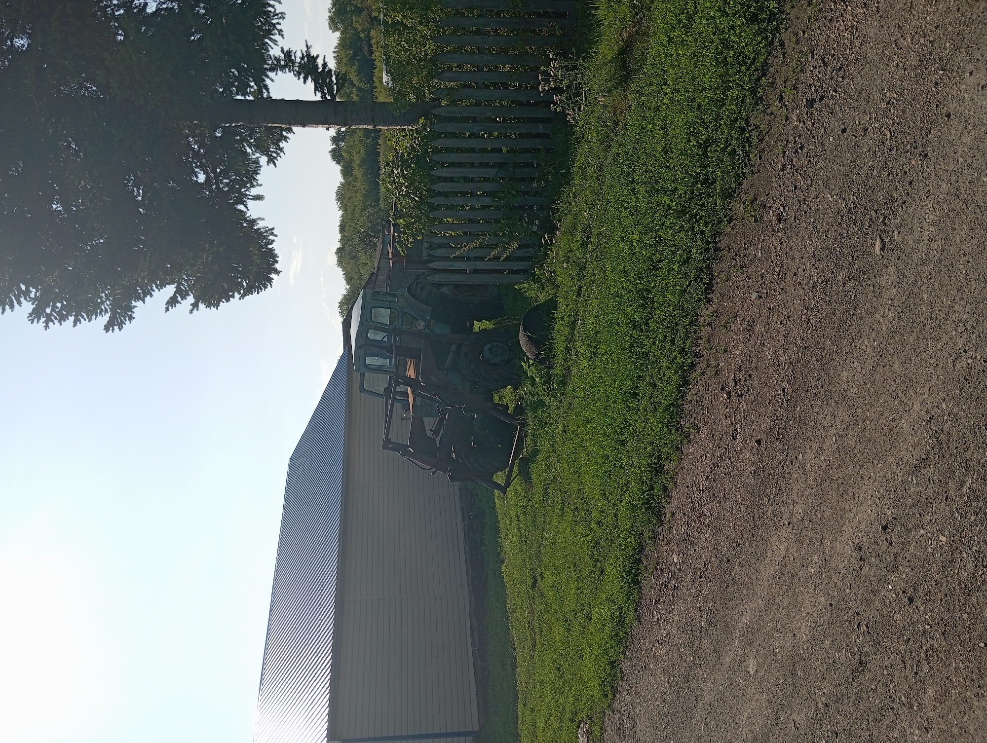 The image depicts a rural scene in Novo-Kopylovo, Russia, on a bright, sunny day. In the foreground, a gravel road runs diagonally across the bottom of the frame. To the left of the road is a patch of vibrant green grass.
Dominating the mid-ground is a green tractor with a front-end loader. It is positioned on the grass, partially obscured by a wooden fence. The fence, made of vertical wooden planks, runs along the right side of the image and extends into the background. Behind the tractor and fence, lush green foliage and trees are visible, indicating a natural environment.
To the left of the tractor and fence, a large, modern building stands. It features a light-colored, horizontal siding and a dark, corrugated metal roof. A large, mature tree with dense foliage is situated behind the tractor and to the right of the building, casting a shadow. The sky above is clear and a pale blue. No people are present in the image.