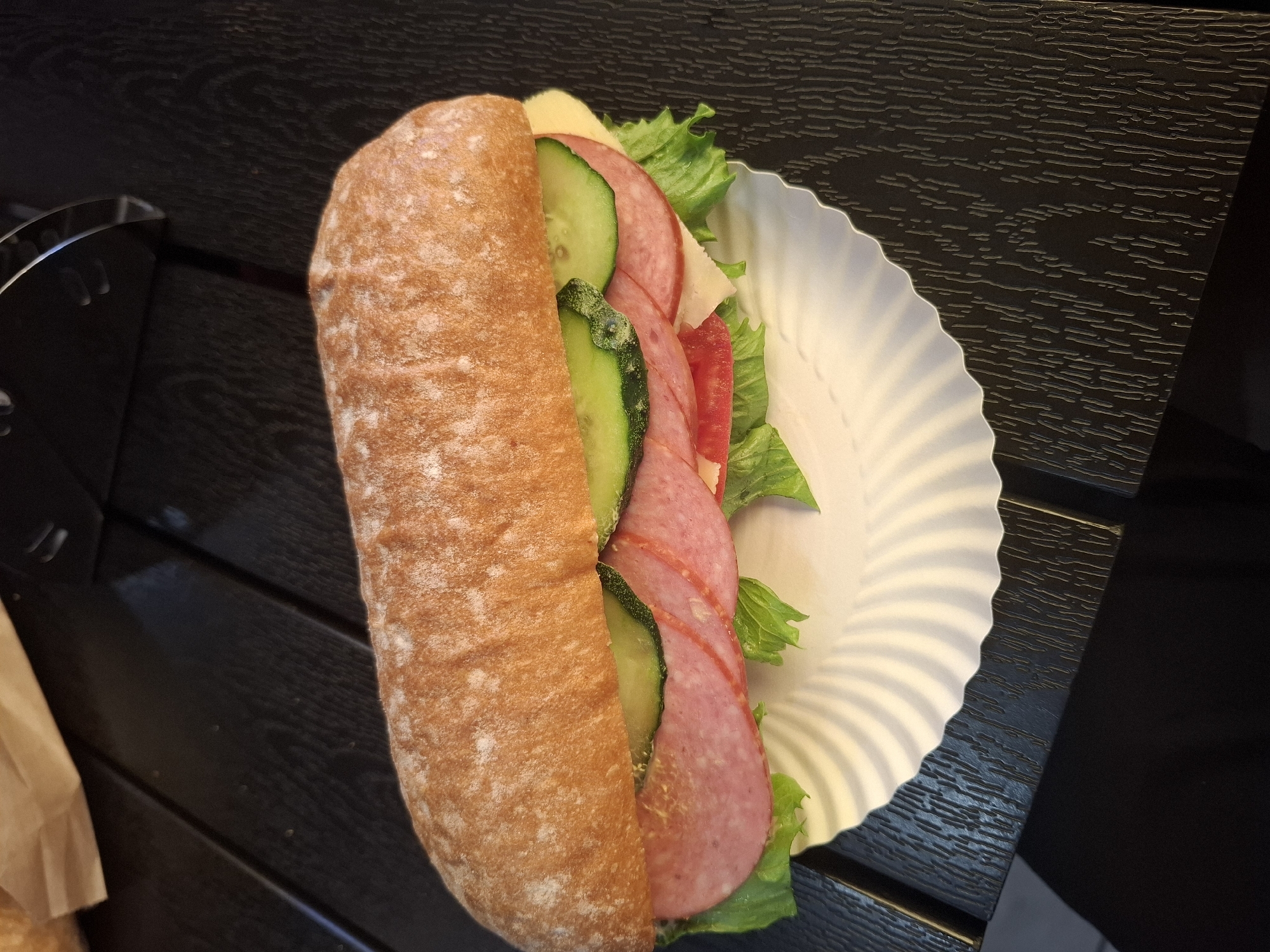 The image shows a close-up, high-angle shot of a sandwich. The sandwich is long and appears to be made with a ciabatta roll, split lengthwise. It is filled with layers of lettuce, salami slices, cucumber slices, tomato slices, and cheese. The sandwich is resting on a white, fluted paper plate, which is placed on a dark, textured wooden surface. A crumpled paper bag is visible in the upper left corner of the image. The lighting suggests an indoor setting, possibly a cafe or restaurant. The scene is devoid of people and any specific geographical markers that would place it in Dubăsarii Vechi, Moldova.