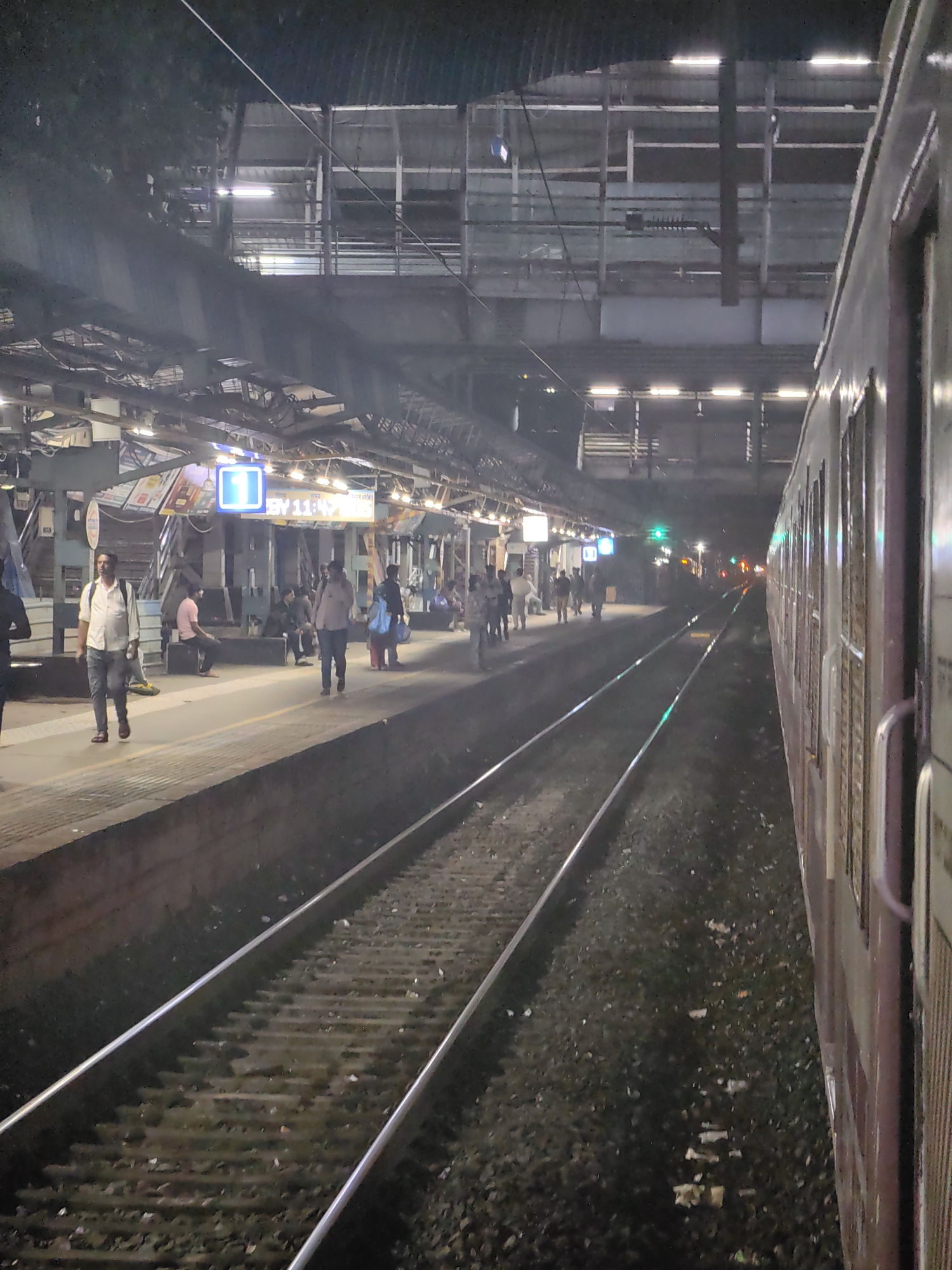 The image depicts a busy train station platform at night, likely in Mumbai, India, given the context of trains and the general atmosphere. A train is visible on the right side of the frame, its carriages painted in muted colors. On the platform to the left, several individuals are present. Some are walking, while others are seated or standing. A man in a white shirt and jeans is walking towards the viewer, carrying a backpack. Another person is on the phone. The platform is illuminated by overhead lights, and a large digital display showing the number "1" and a time (BY 11:47) is prominently positioned. Train tracks run parallel to the platform, extending into the distance. The environment appears to be an urban railway station with the typical infrastructure of platforms, tracks, and overhead coverings.