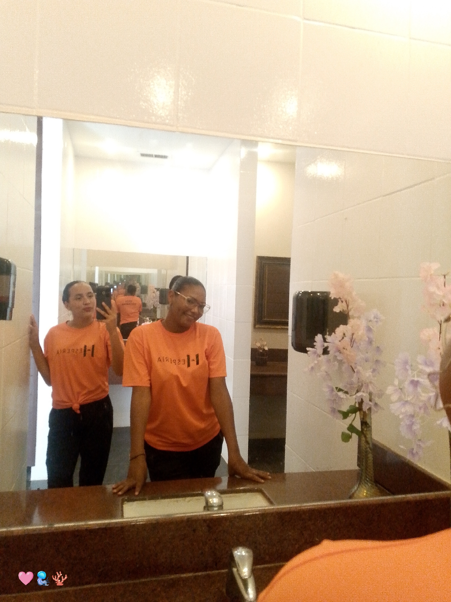 In a restroom at an unknown establishment in Pedro González, Venezuela, two individuals are visible in a mirror. The person on the left, with dark hair pulled back, is holding a black smartphone and taking a selfie. They are wearing an orange t-shirt tied at the waist and black pants. To their right, another person with glasses is also looking towards the mirror, wearing a similar orange t-shirt and black pants. The restroom features white tiled walls, a brown countertop with a sink and faucet, and a decorative vase with light purple and pink faux flowers. In the reflection, another person wearing an orange shirt is visible further back in the restroom, as is a framed artwork on the wall.