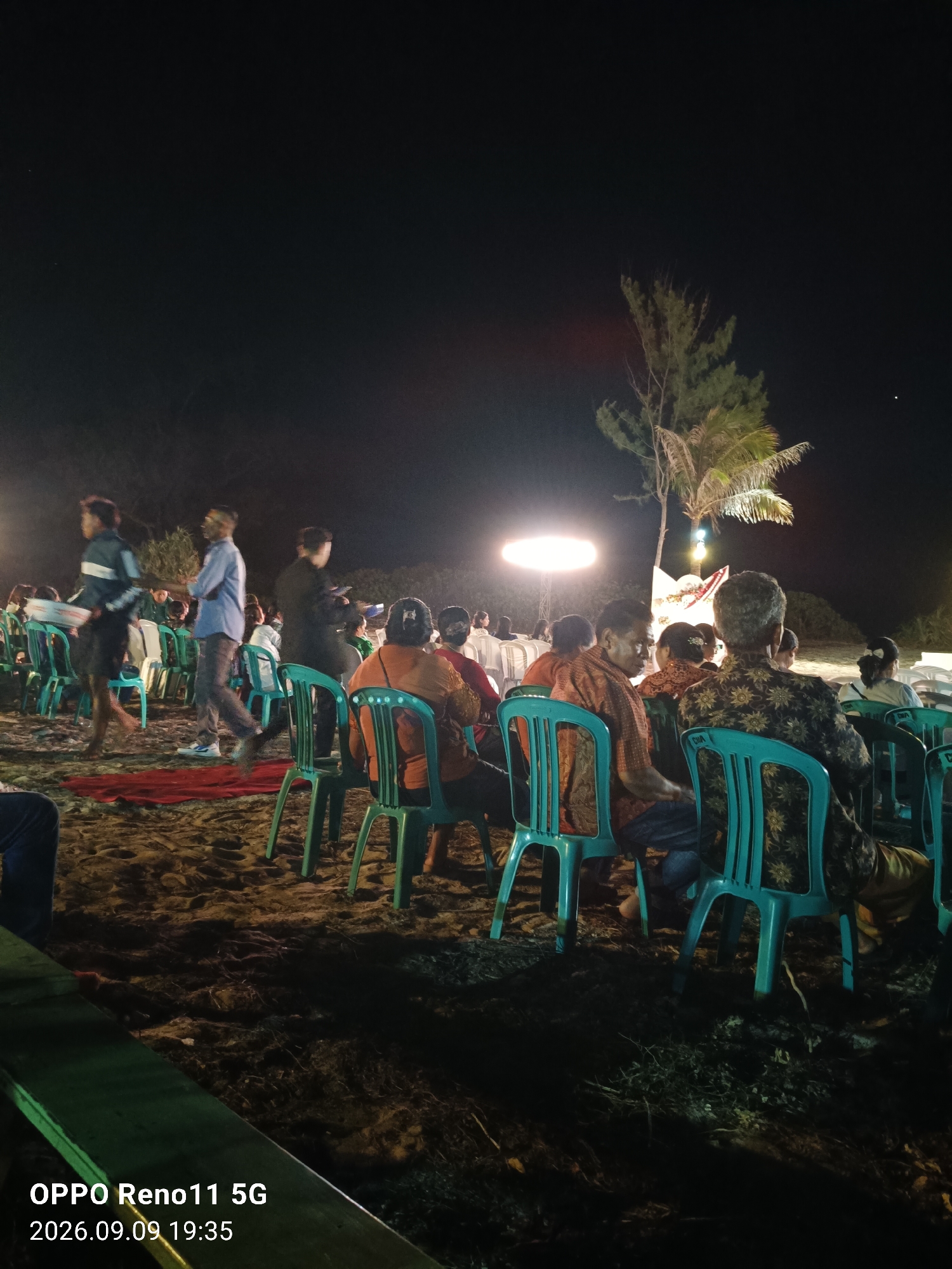 The image depicts an outdoor gathering at night, likely on a beach given the sandy ground and a distant tree that resembles a palm. Several people are seated on blue plastic chairs, facing a central area where a bright light source is visible. Some individuals are standing and walking, suggesting an event is in progress. The setting appears to be in Otan, Indonesia, as indicated by the text overlay which includes "OPPO Reno11 5G" and a date and time stamp of "2026.09.09 19:35". The lighting is artificial, with strong spotlights illuminating the central area and casting shadows. The overall atmosphere appears to be of a community event or performance.