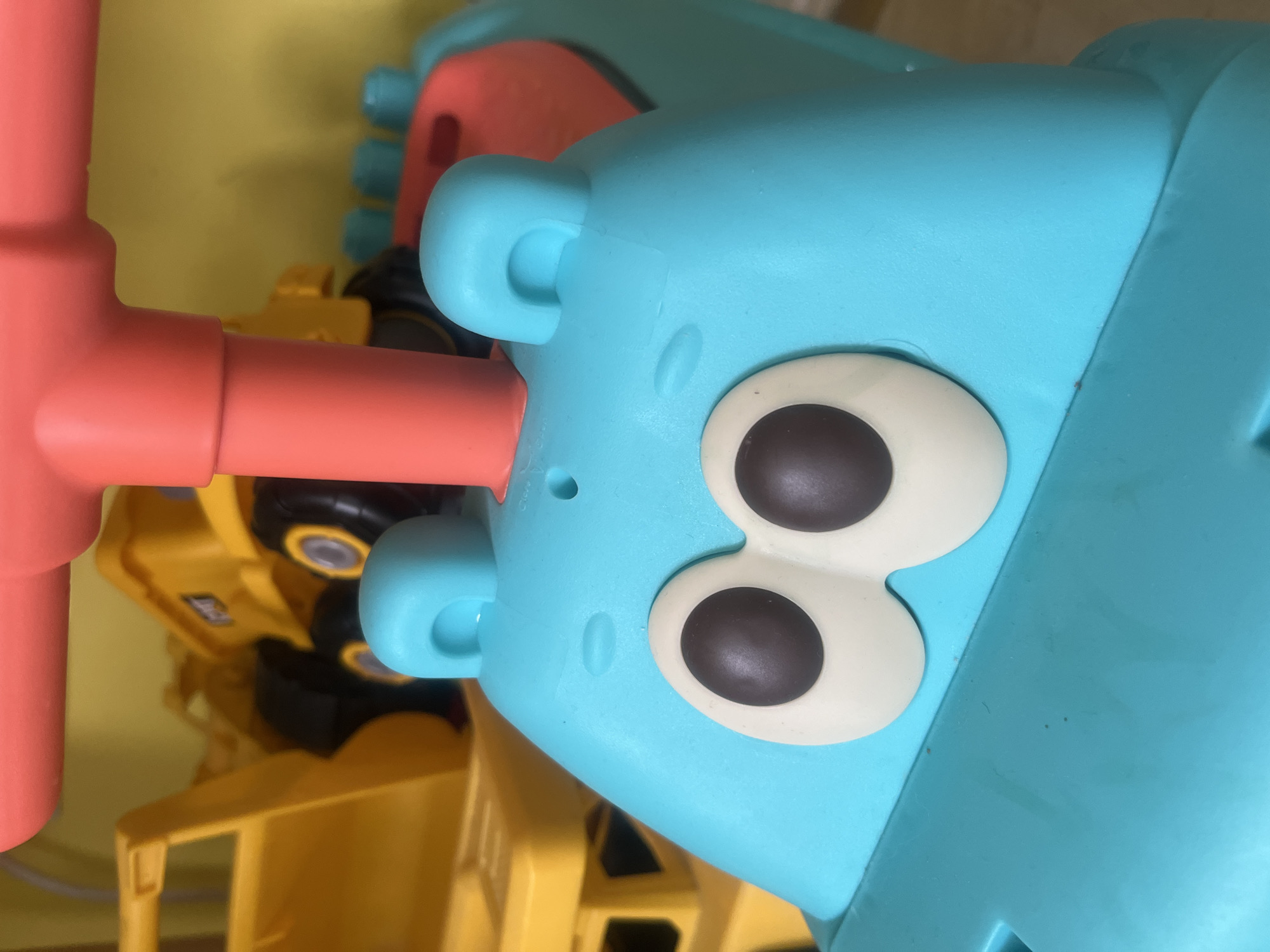 This is a close-up, eye-level, indoor photograph of a blue ride-on toy in the shape of an animal, possibly a hippopotamus or frog, with large cartoon eyes. The toy is turquoise blue with oversized dark gray eyes outlined in white. It has two rounded turquoise ears on top. A bright coral-colored handle extends upwards from the toy's head. In the background, out of focus, are yellow toy construction vehicles. The background wall is a solid yellow color. The image does not contain any information that would allow for the identification of its location in the city of Onokivtsi, Ukraine.