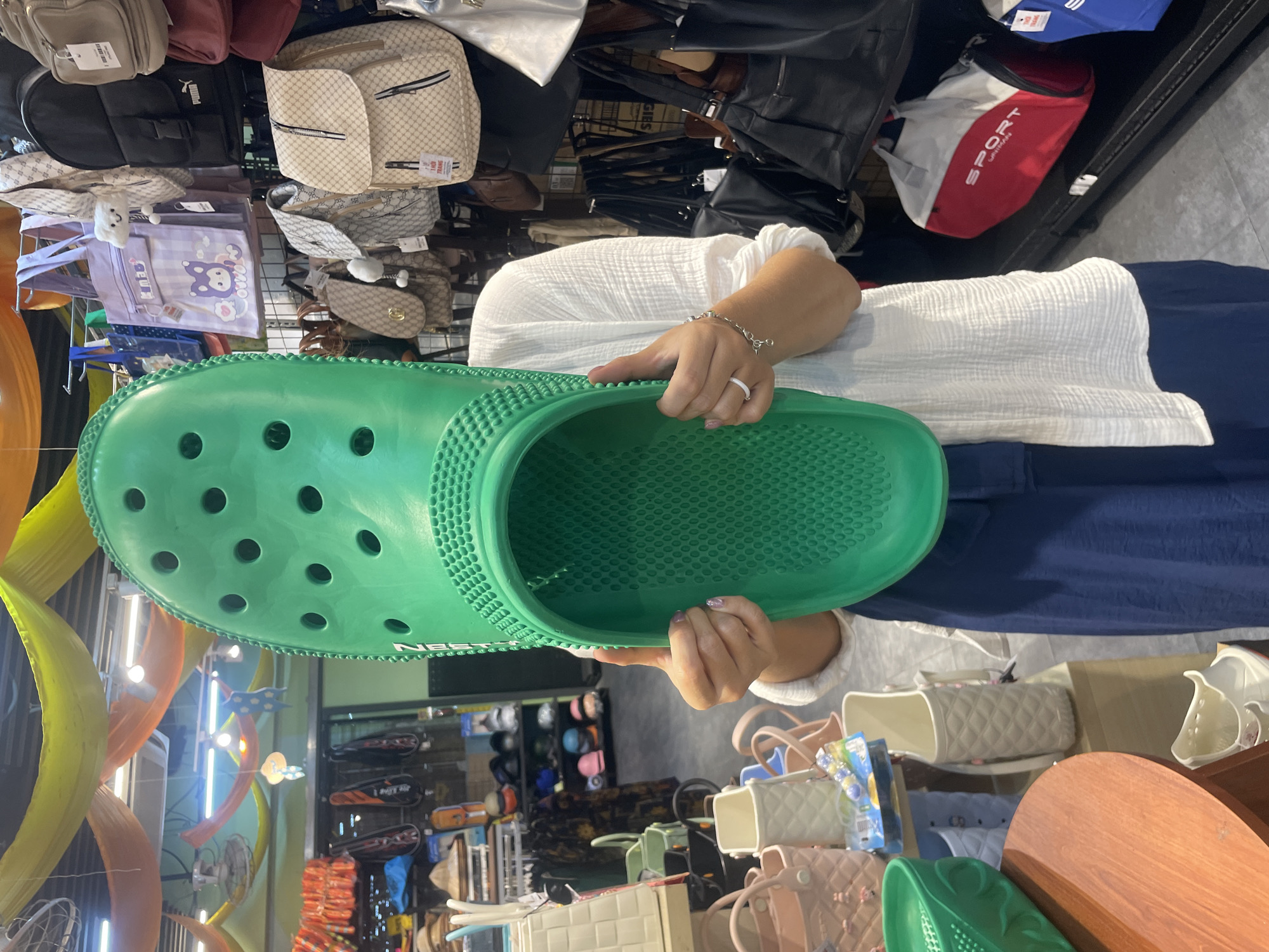 A person wearing a white top and dark blue skirt is holding up a very large green clog inside a shop in Bac Nha Trang Ward, Vietnam. Bags and other merchandise hang on racks behind the person, and other products are displayed on tables below.