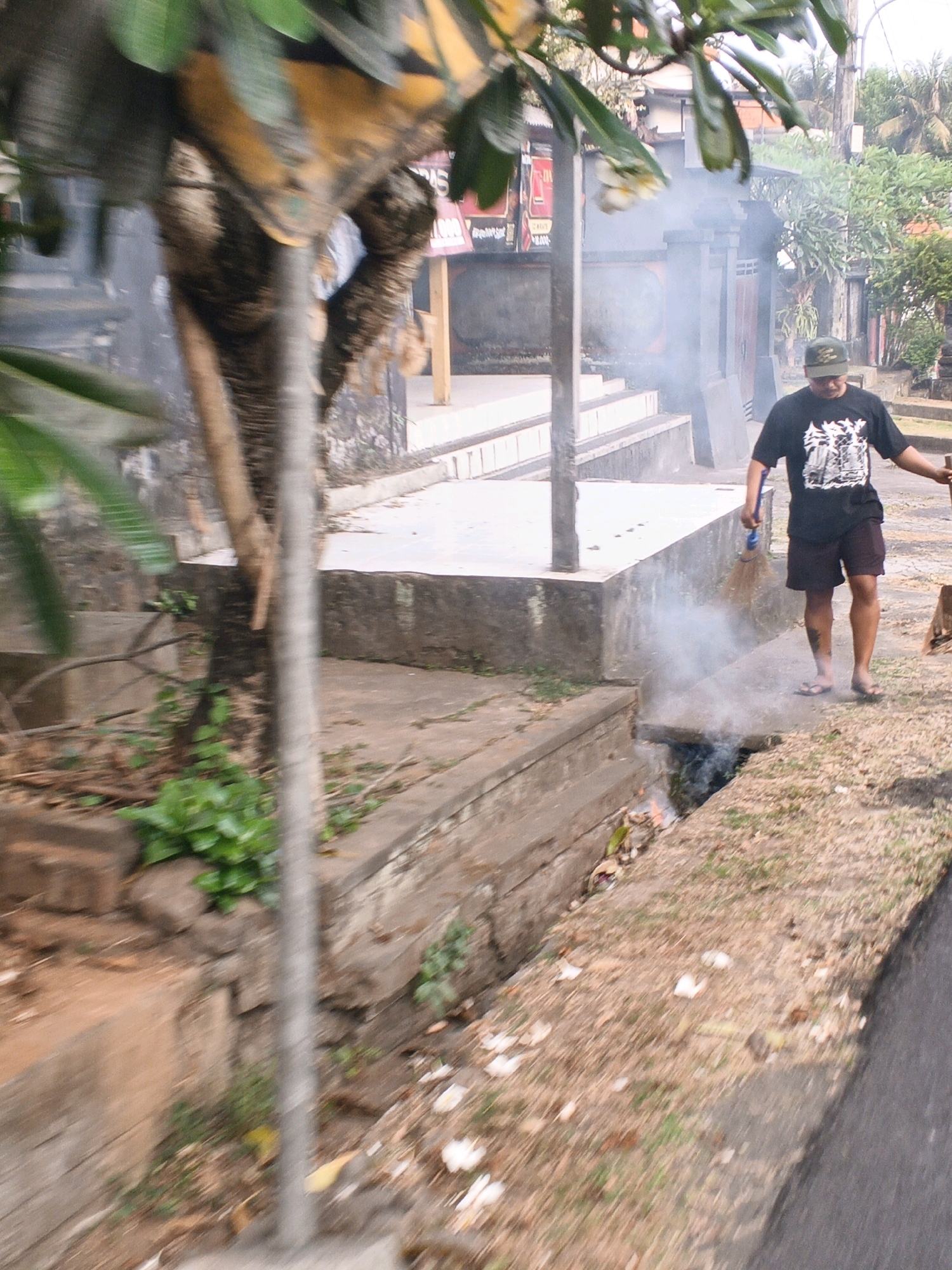A man wearing a dark t-shirt with a white graphic, dark shorts, a dark baseball cap, and flip-flops is sweeping a small fire in a drainage ditch with a broom. Smoke is rising from the ditch. The man is standing on a dirt path next to a concrete drainage system. In the background, there is a building with a dark door and a gray wall. There are also trees and plants. The image appears to be taken in Sukawati, Indonesia.