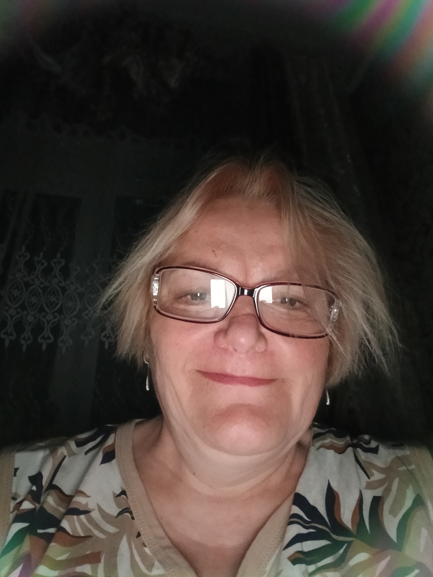 A medium shot depicts a smiling, mature woman with short, layered blonde hair and glasses. She is wearing a patterned top, and small, dangling earrings are visible. The background is dark, suggesting an indoor setting, possibly near a window with patterned curtains. There is nothing in the image that suggests this photo was taken in Красный Маяк, Russia.