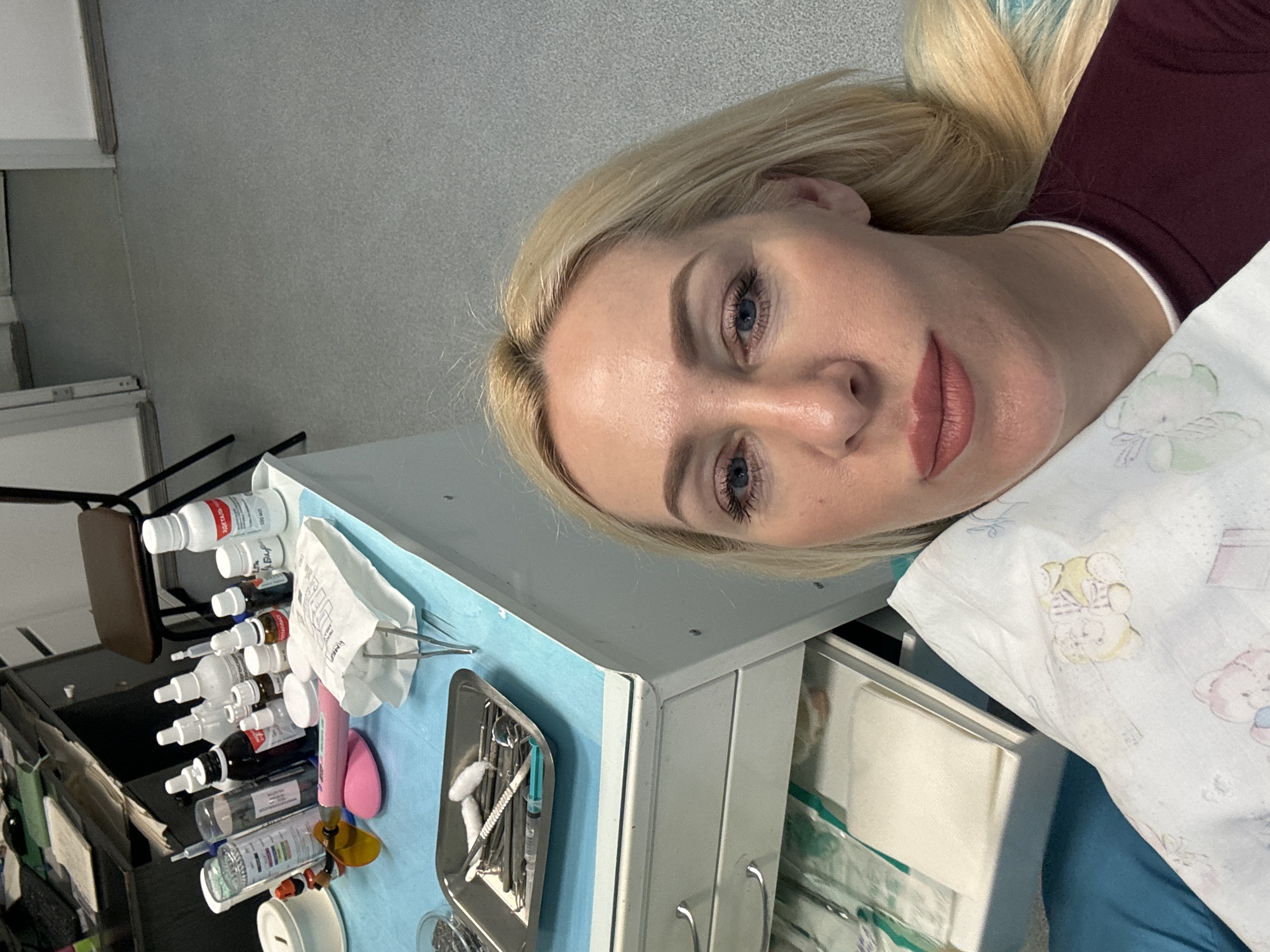 The image shows a blonde woman with blue eyes and fair skin, wearing a maroon shirt. She is lying down and holding a white cloth with teddy bear prints. Her face is partially visible, with makeup on her eyes and lips. In the background, a desk is covered with various medical supplies, including bottles of liquids, syringes, instruments on a tray, and a paper bag containing medical tools. The overall setting appears to be a medical or dental clinic. The floor is grey and speckled, and a stool is visible in the background. The scene does not provide any specific clues about the location in Saint Petersburg, Russia.