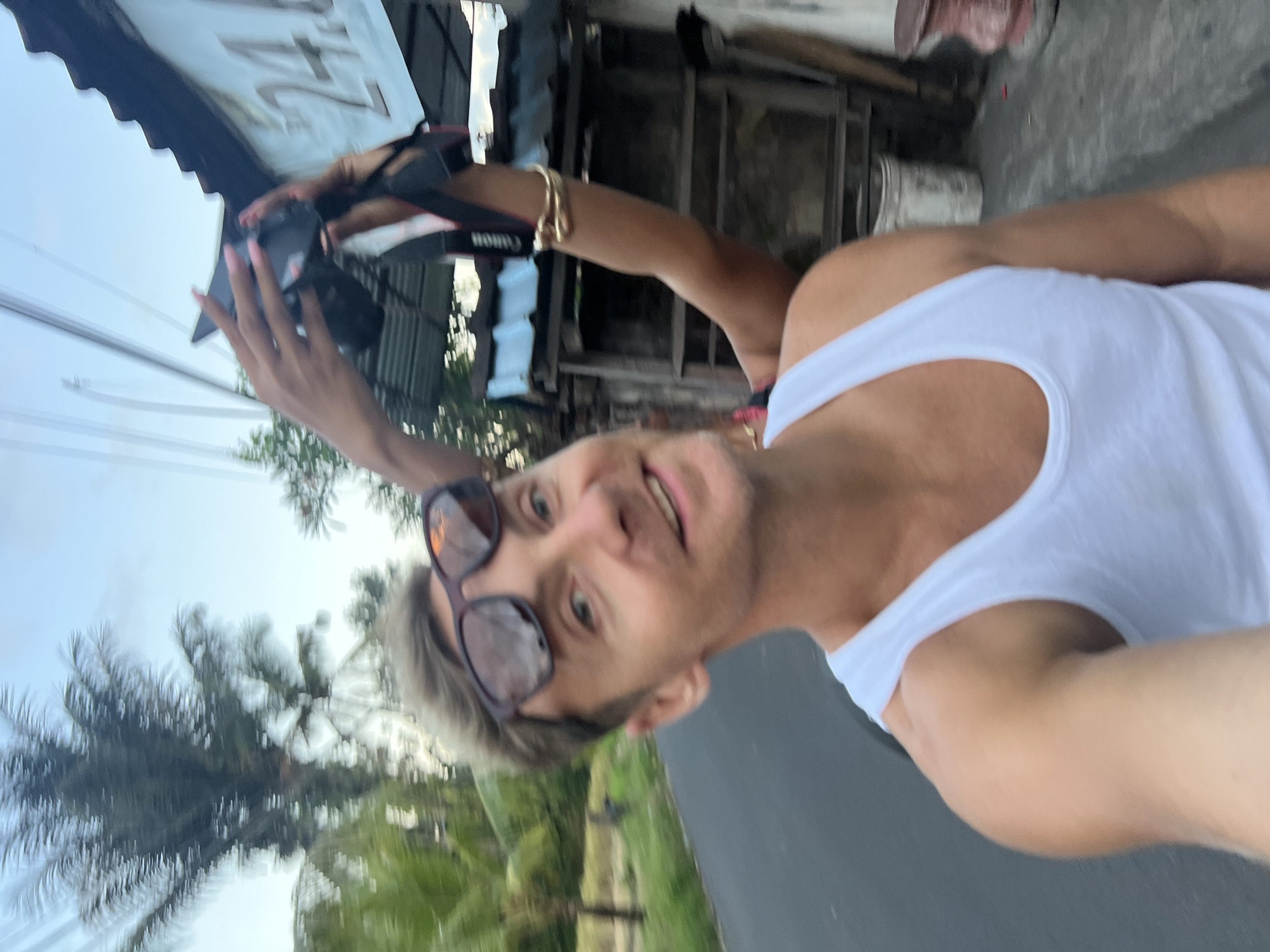 A man is posing for a selfie on a street in Sukawati, Indonesia. He is wearing a white tank top and sunglasses. In the background, a person is holding a Canon camera. Palm trees and buildings are also visible.