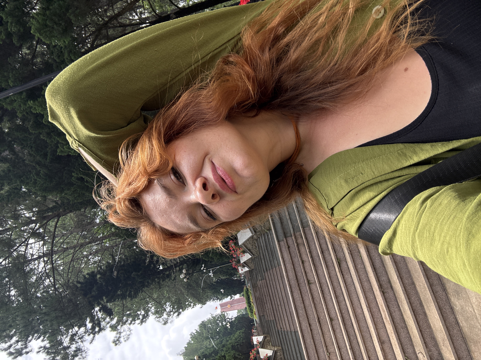 The image is a close-up, selfie-style shot of a woman with long, wavy, auburn hair. She is wearing a green jacket over a black top, and a thin, brown necklace. Her left arm is raised, with her hand resting near her head, and she is looking directly at the camera with a slight smile.
The background appears to be an outdoor setting, possibly a park or memorial site, characterized by a wide set of stone stairs leading upwards. Along the sides of the stairs are planters with red flowers and white decorative elements. In the distance, a tall, slender red and white structure is visible, topped with a flag. The area is surrounded by trees, many of which appear to be pine trees, suggesting a cooler climate or elevation. Streetlights are also visible along the walkway. The sky is overcast.
This scene is likely located at the Cam Ly waterfall or the surrounding area in Đà Lạt, Vietnam, given the distinctive stone stairs, planters, and the memorial structure often seen in that region.