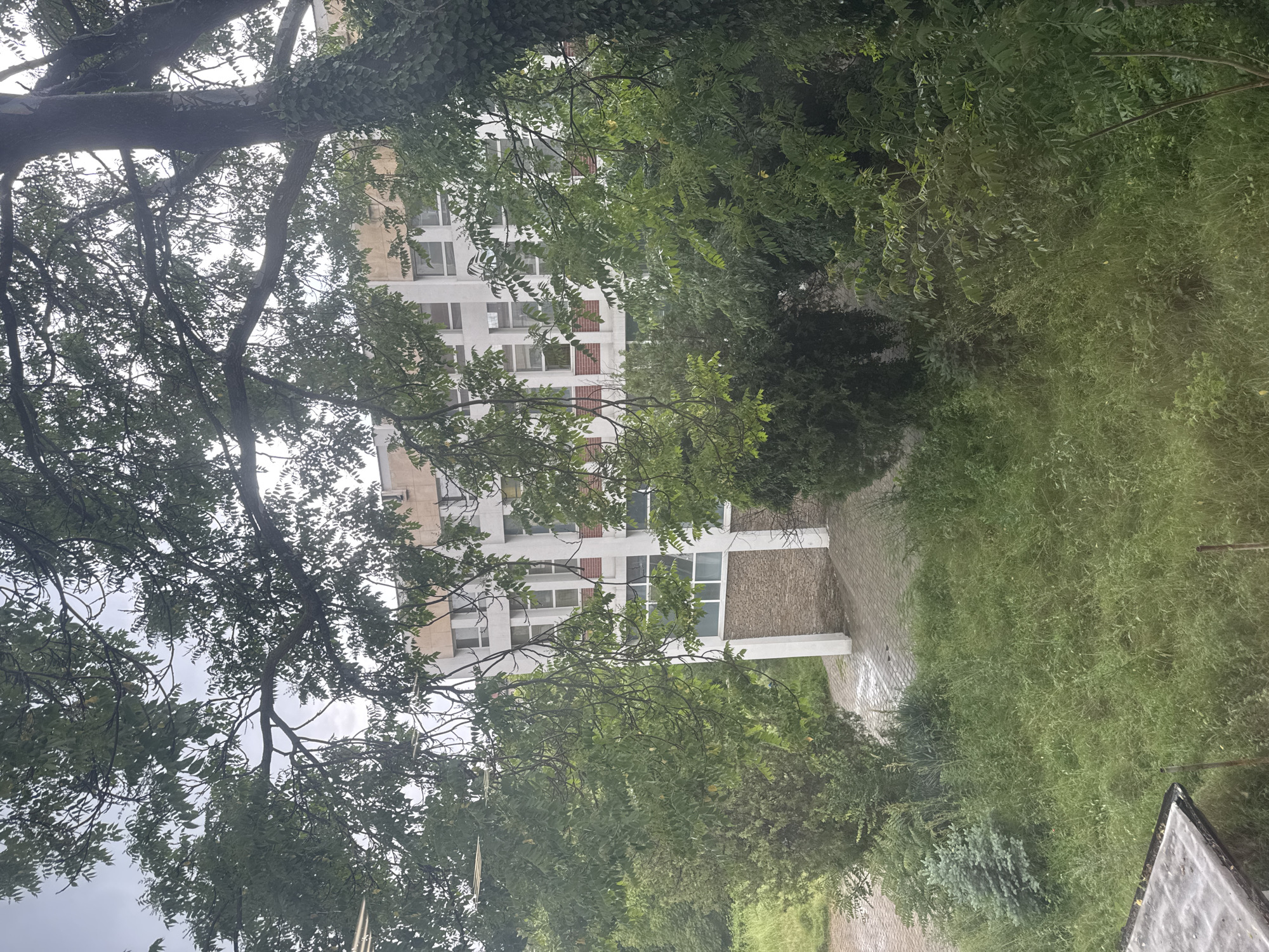 The image shows a partially obscured view of a building with white walls and multiple windows, some of which have red accents. The building appears to be surrounded by trees and vegetation. A paved area is visible in front of the building. The location is Unknown, Unknown.