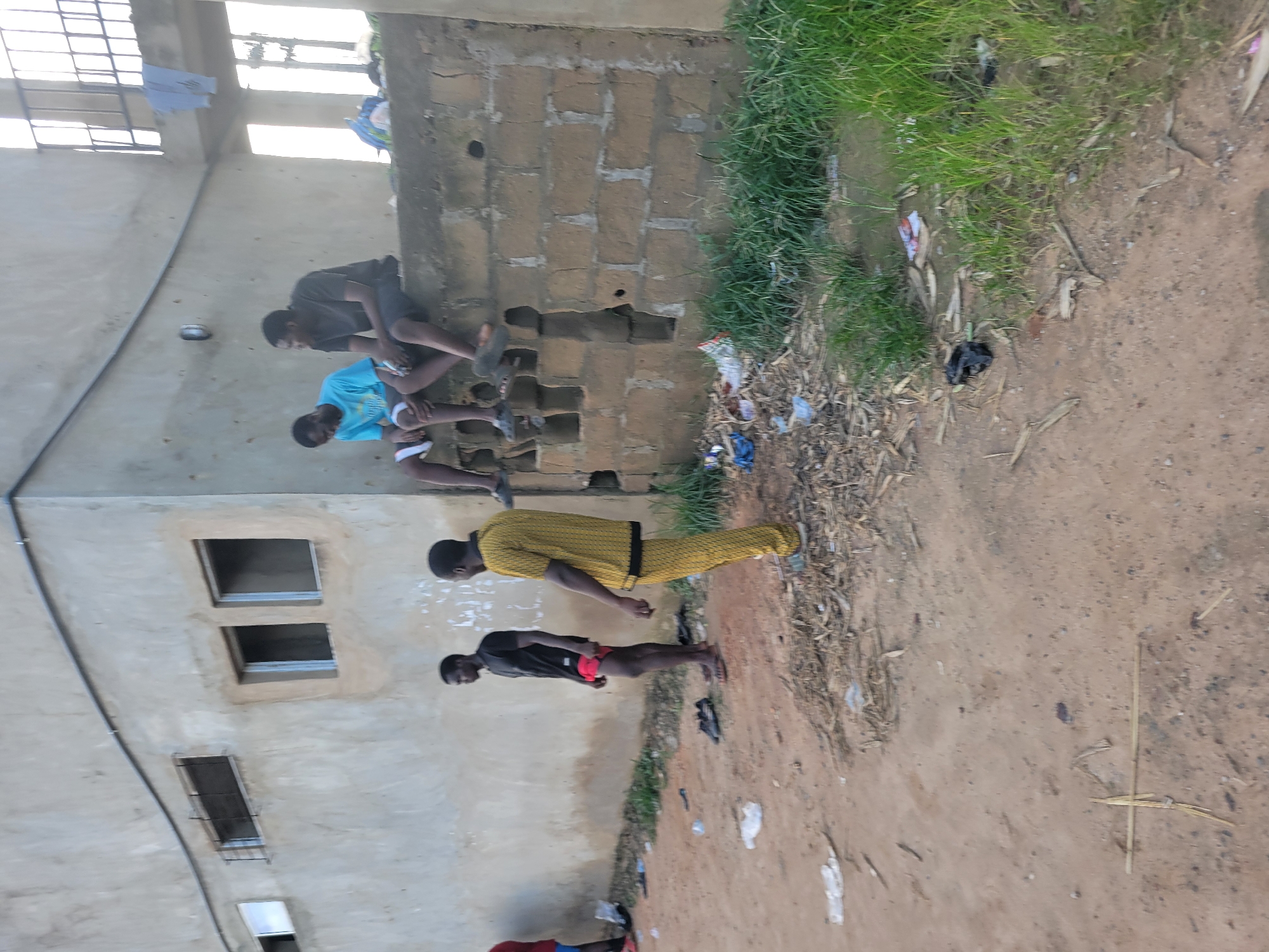 The image shows a group of young men gathered outside a building in what appears to be a rural or semi-urban setting. The building has a rough, brick-like texture and features several small windows. One man is wearing a yellow, patterned shirt and dark pants, standing with his hands clasped behind his back. Another man in a black t-shirt and red shorts stands to his right, looking down. Two other men are seated on a ledge or low wall, one in a dark shirt and shorts, the other in a bright blue t-shirt and shorts. The ground is dirt, with some sparse vegetation and debris scattered around. The lighting suggests it is daytime, possibly late afternoon, with soft shadows. The overall atmosphere appears casual and relaxed, with no obvious signs of distress or urgency. The presence of laundry hanging in an open area above the seated men might suggest a residential or communal living space. The location context provided is Egbagu, Nigeria. No visible text is present in the image.