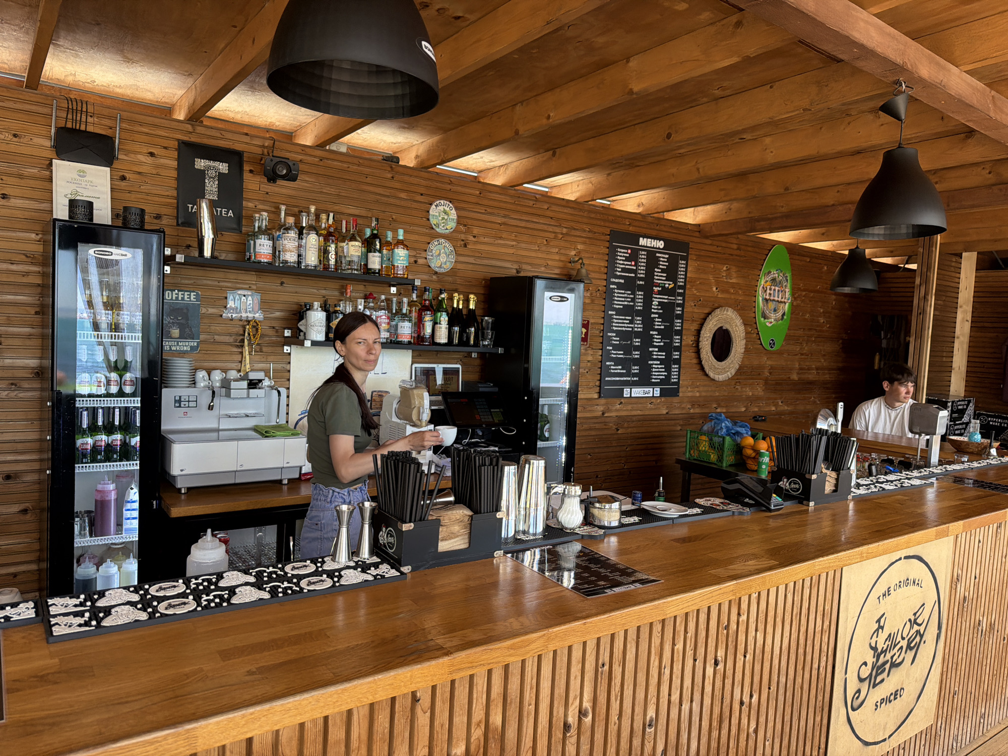 The image depicts the interior of an outdoor bar, likely in Burgas, Bulgaria. A woman stands behind the wooden bar, preparing a drink. Behind her, there is a shelf stocked with various liquor bottles, a coffee machine, and a refrigerator filled with beverages. A menu board hangs on the wall. The bar itself is made of wood and displays the "Sailor Jerry Spiced" logo on its front. A young man sits behind the bar on the right side of the frame. Black pendant lights hang from the wooden ceiling. There are bar accessories such as straws and jiggers. The bar appears to be well-stocked and organized.