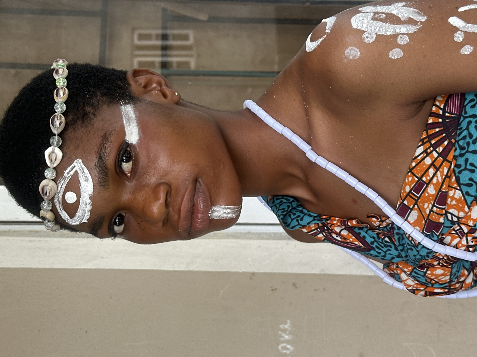 A young Black woman with short, dark hair stands against a neutral-toned wall, looking directly at the camera. She has white face paint, including an abstract eye shape on her forehead, a stripe under her left eye, and a vertical stripe on her chin. She wears a headband adorned with shells and beads, and a light blue beaded necklace. Her outfit is a colorful, patterned fabric with orange, teal, and maroon accents, draped over her shoulder. White paint is also visible on her right shoulder and upper arm, in abstract circular and linear patterns. The background includes a doorway with glass panes and a portion of a light-colored wall with faint markings. The scene is set in Badagry, Nigeria.