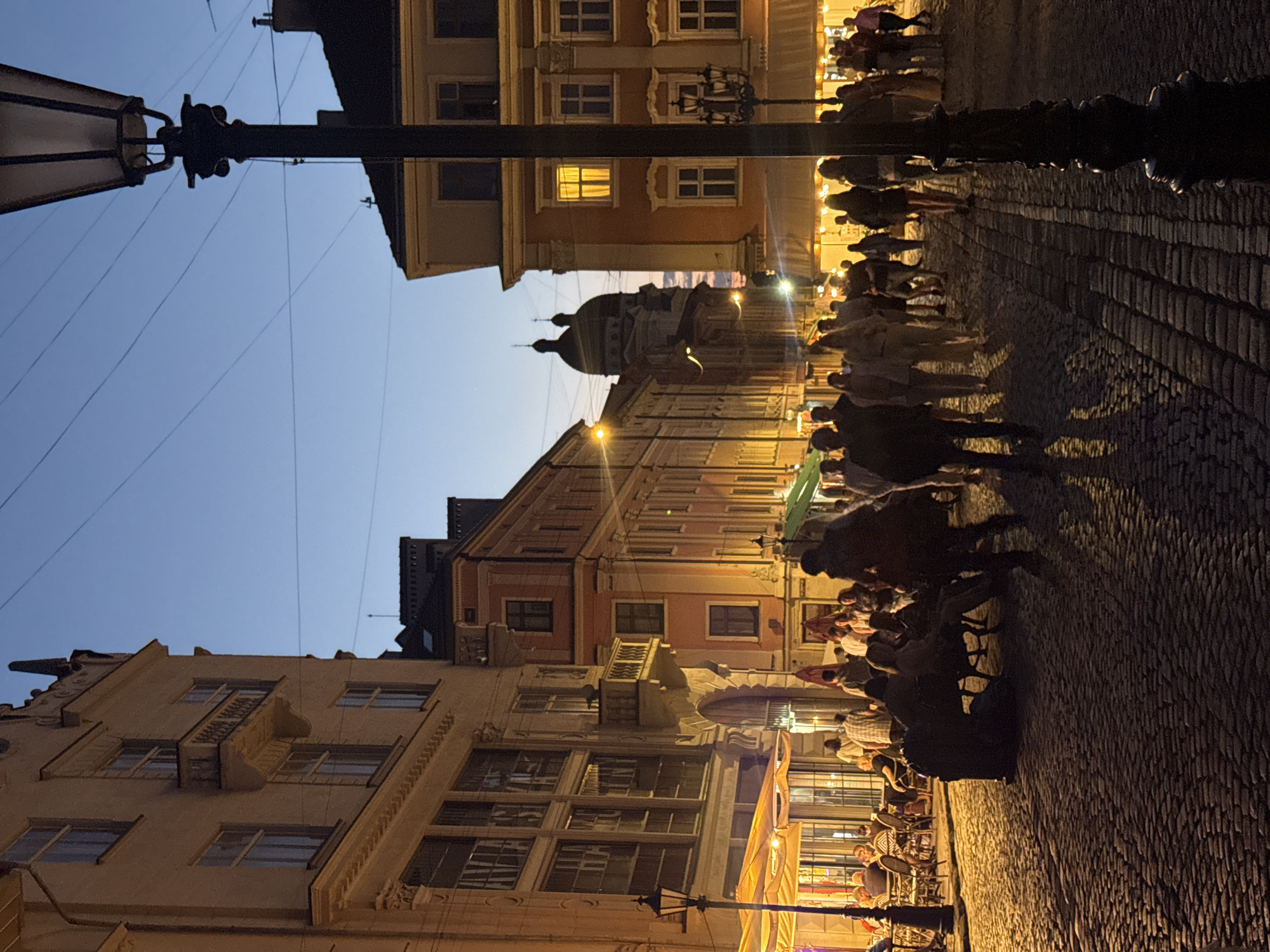 An outdoor, eye-level, wide-angle shot captures a cobblestone street in Lviv, Ukraine, at dusk. A lamppost stands in the foreground, its black, ornate pole casting a shadow on the textured ground. Buildings line both sides of the street, their facades illuminated by warm, artificial light from windows and street lamps.
On the left, a multi-story building features a light-colored facade with ornate architectural details and a small balcony. A yellow awning extends from a ground-level establishment, with patrons seated at tables under its cover, suggesting a cafe or restaurant.
The right side of the street showcases buildings with orange and pink facades, also with multiple windows. Further down the street, a large domed structure with two cupolas is visible against the twilight sky.
A crowd of people is present, walking along the street and gathered in small groups. Their figures are mostly silhouetted against the illuminated background, with some casting distinct shadows on the cobblestones. The atmosphere appears lively, with a sense of evening activity. The sky transitions from a deep blue at the top to lighter hues near the horizon, indicating the end of daylight.