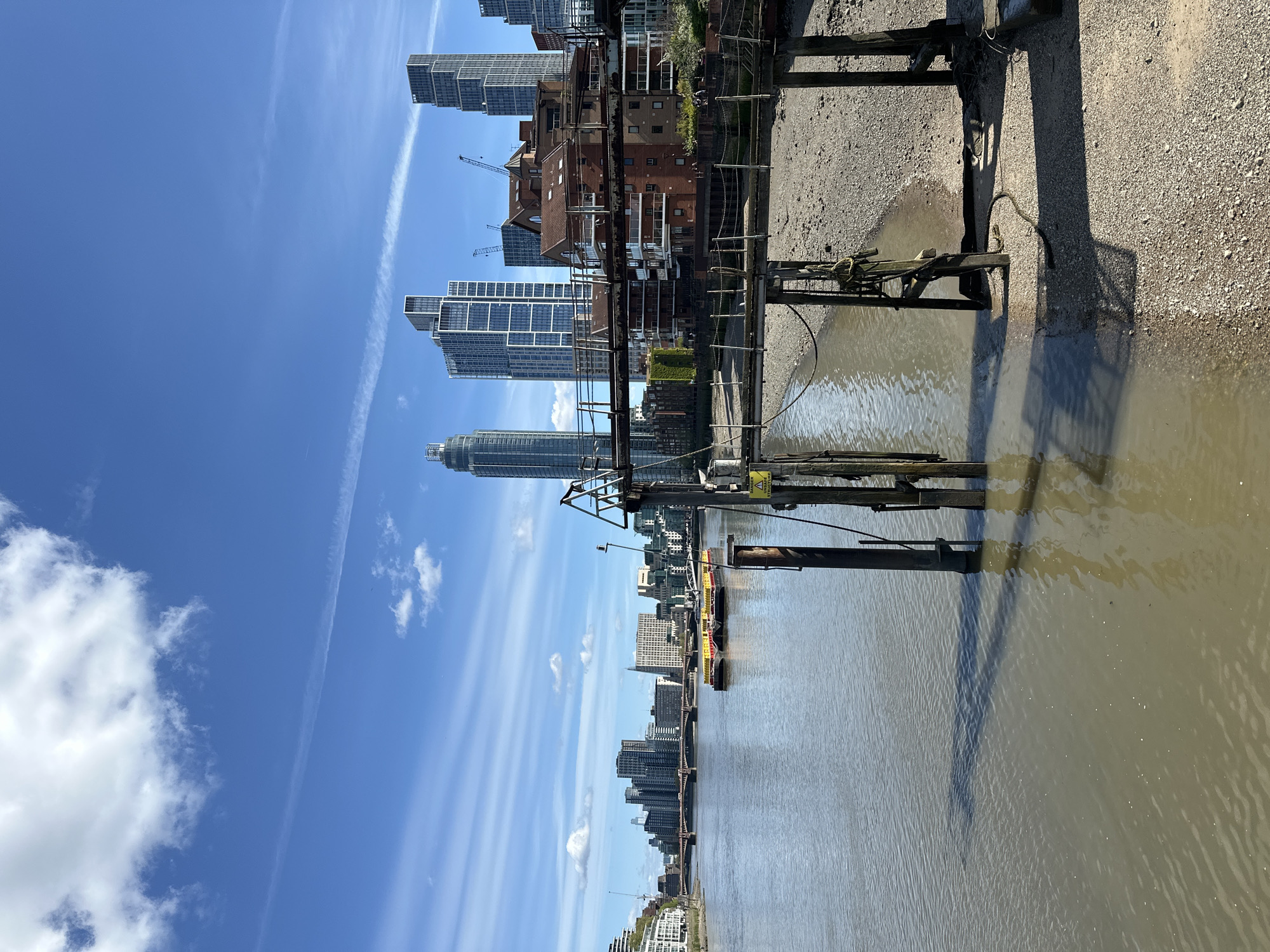 The image depicts a waterfront scene in London, United Kingdom. The perspective is from a low angle, close to the water's edge, possibly from a pier or similar structure. In the foreground, weathered wooden pilings and parts of a decaying structure extend into the murky water. The water is a light brown color, typical of the Thames Estuary.
In the middle ground, there is a view of a city skyline. Modern skyscrapers are visible, mixed with older brick buildings. A crane signifies ongoing construction. There are barges or workboats on the water in the middle distance.
The background is a bright blue sky with scattered clouds. The lighting suggests a sunny day. The overall impression is one of urban development juxtaposed against the natural element of the river and the decay of older structures.