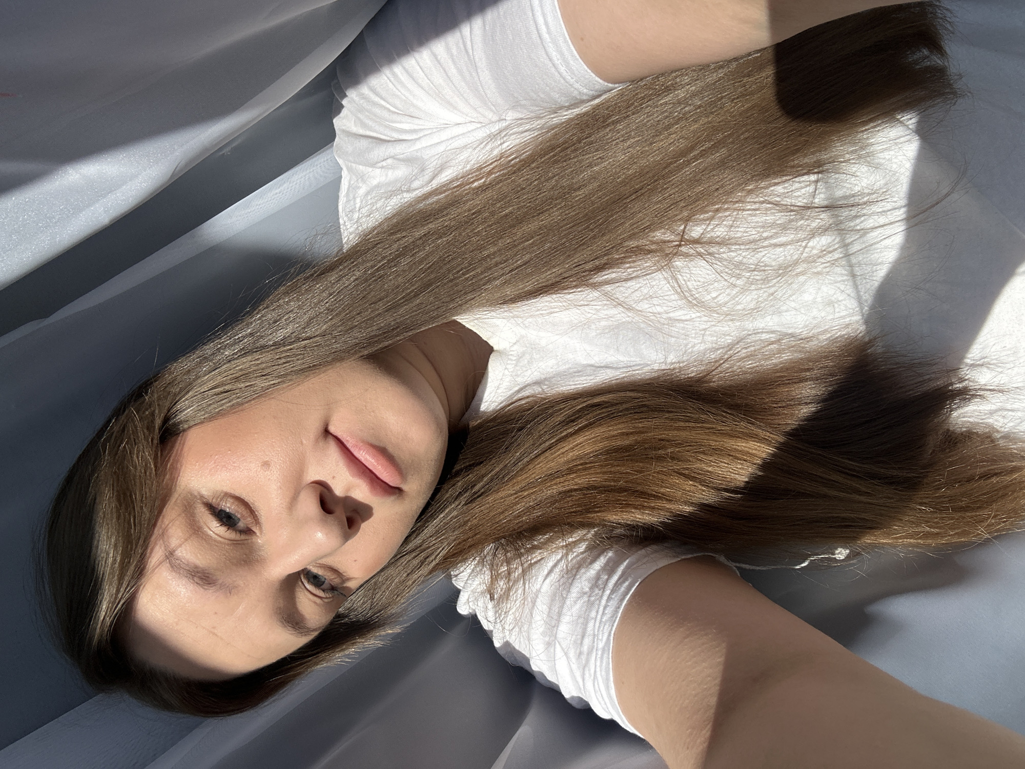 A woman with long, straight brown hair lies on a gray surface in Ольховка, Russia. She is wearing a white t-shirt and her face is partially in shadow, her lips slightly parted. The background features a soft, gray fabric draping around her.