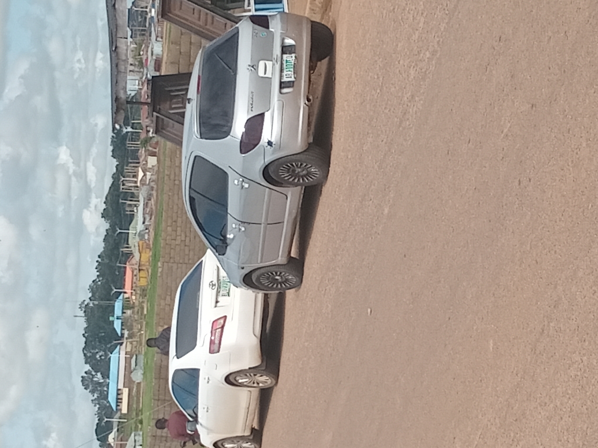 In Bukuru, Nigeria, two parked cars, a white Toyota Camry and a silver Peugeot 307, are visible on a dirt road under a cloudy sky. The Toyota Camry is to the left, and the Peugeot 307 is to the right. Two individuals, one wearing a maroon shirt and the other a dark jacket, are standing near the Toyota Camry. In the background, a brick wall and several buildings are present, with trees and construction structures beyond them. The road surface is coarse and dusty.