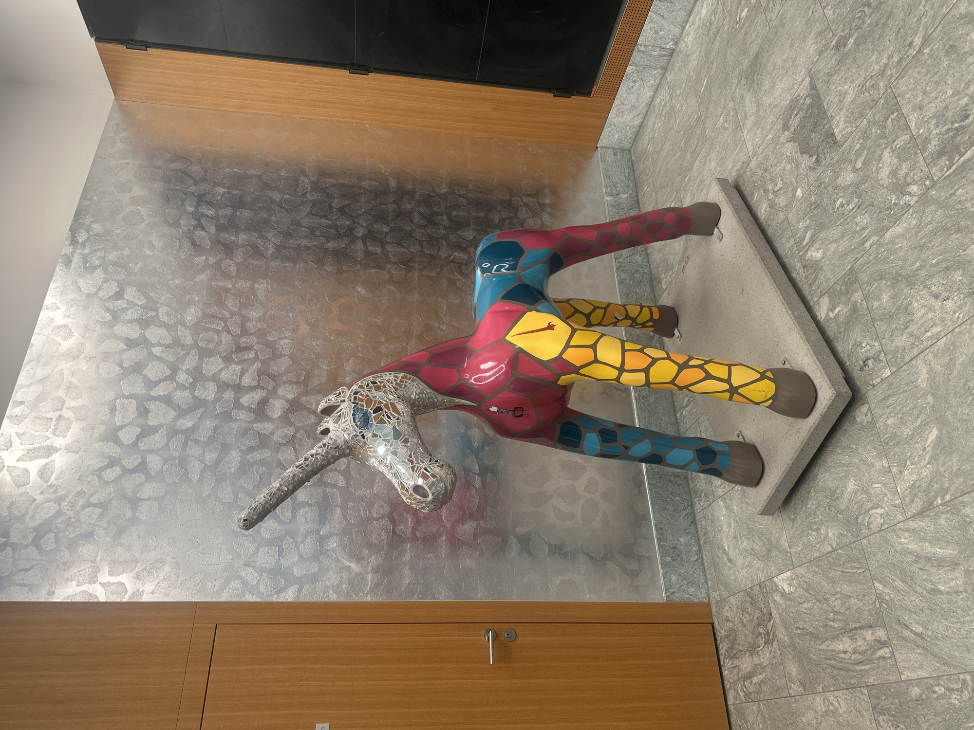 The image depicts a mosaic unicorn sculpture standing on a concrete base indoors. The unicorn's head is covered in a mosaic of small mirror-like tiles, while its body and legs are painted in geometric patterns of red, yellow, blue, and green. The sculpture is positioned against a wall with a textured, metallic-looking silver covering. In the foreground, there is a light brown wooden structure, possibly a cabinet or paneling. The floor is tiled in a light gray stone pattern. The sculpture is located in the city of Dübendorf, Switzerland.