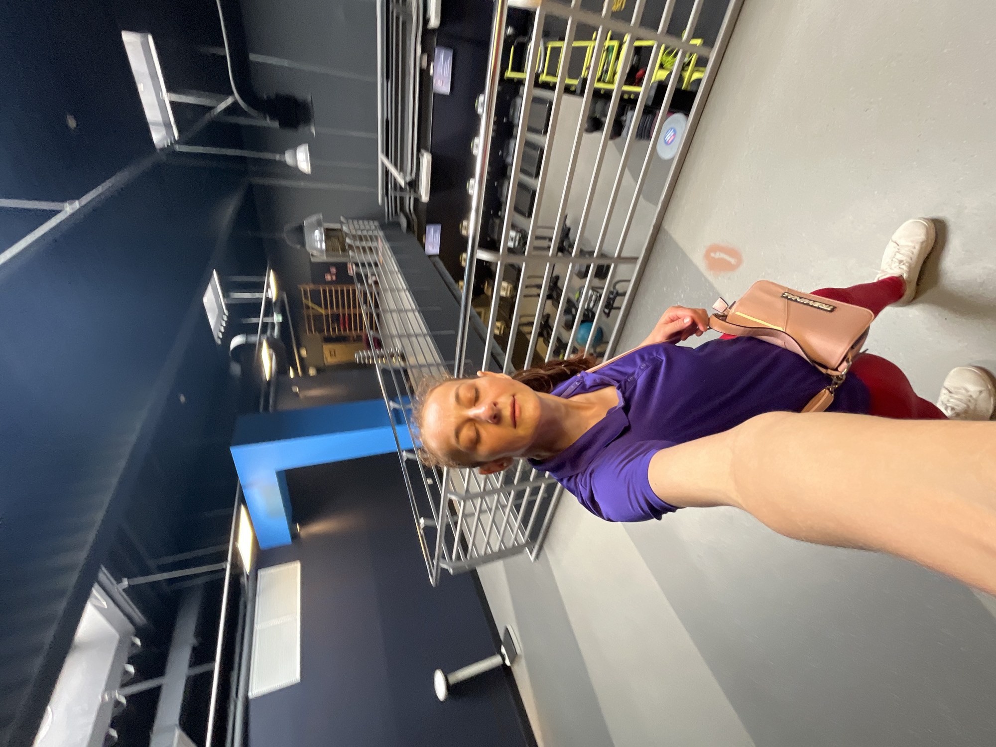 A woman is taking a selfie in what appears to be a gym or fitness facility. She is wearing a purple polo shirt, red leggings, and white sneakers. She is holding a light pink purse in her left hand. Her eyes are closed, and she has a slight smile on her face. The background shows multiple levels of a spacious interior with metal railings, gym equipment like treadmills and weight machines, and industrial-style lighting. A large, bright blue architectural element is visible on the left side of the image. The overall lighting is dim, with artificial lights illuminating the space.