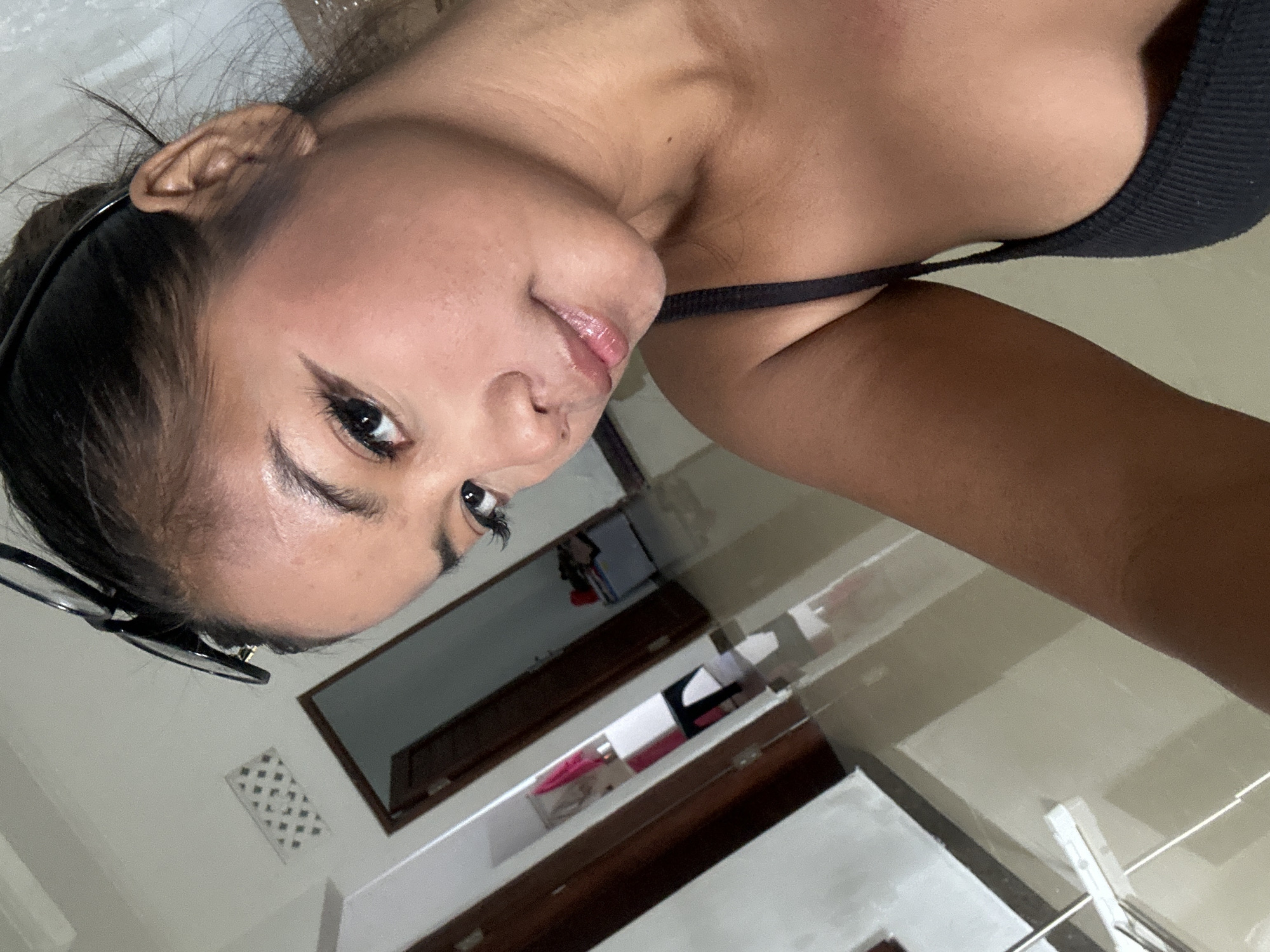 A selfie is taken from a low angle, showing a woman's face and upper body. She has dark hair pulled back, with sunglasses resting on top of her head. She is wearing a black tank top. Her skin is tanned, and she has makeup on, including eyeliner and lip gloss. The background appears to be an indoor hallway, possibly in a home or hotel, with tiled floors and several doors visible. The location is identified as Sukawati, Indonesia.