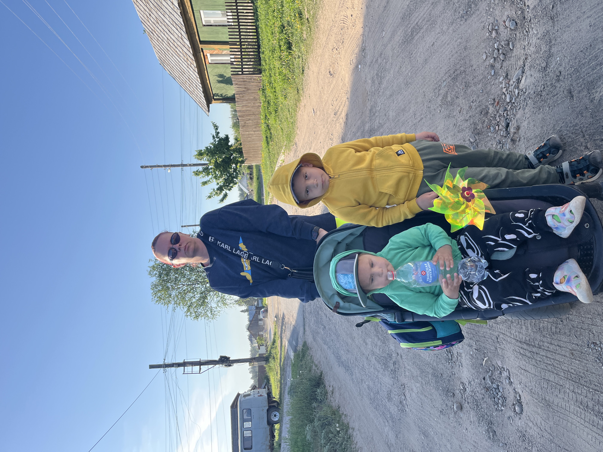 A man, wearing sunglasses and a dark sweatshirt, stands on a gravel road in a location identifiable as the city of Chaikovsky, Russia. Beside him is a stroller containing two children. The child in the stroller's seat wears a green shirt and drinks from a plastic water bottle labeled in Russian. The older child, wearing a yellow hooded sweatshirt and green pants, lies in the stroller. They hold a multi-colored pinwheel. A building with a green exterior and a gray roof is visible in the background, along with power lines and a light-colored vehicle. The sky is blue and clear.