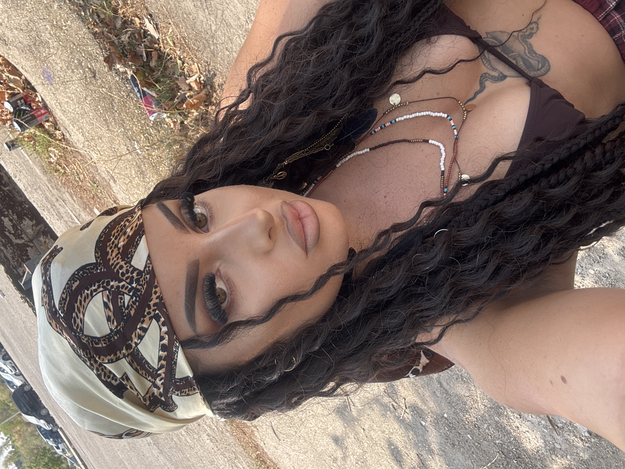 A woman with long, dark, curly hair and dramatic makeup, including false eyelashes and contouring, is shown from the chest up in a selfie. She is wearing a patterned headscarf with snake and leopard print, a dark bikini top, and layered necklaces. A tattoo is visible on her chest. The background suggests an outdoor urban environment, with a paved surface, dry vegetation, discarded energy drink cans, and parked cars in the distance. The lighting indicates it is daytime. The image is likely a casual personal photograph. The scene is set in an open area with signs of neglect and litter.