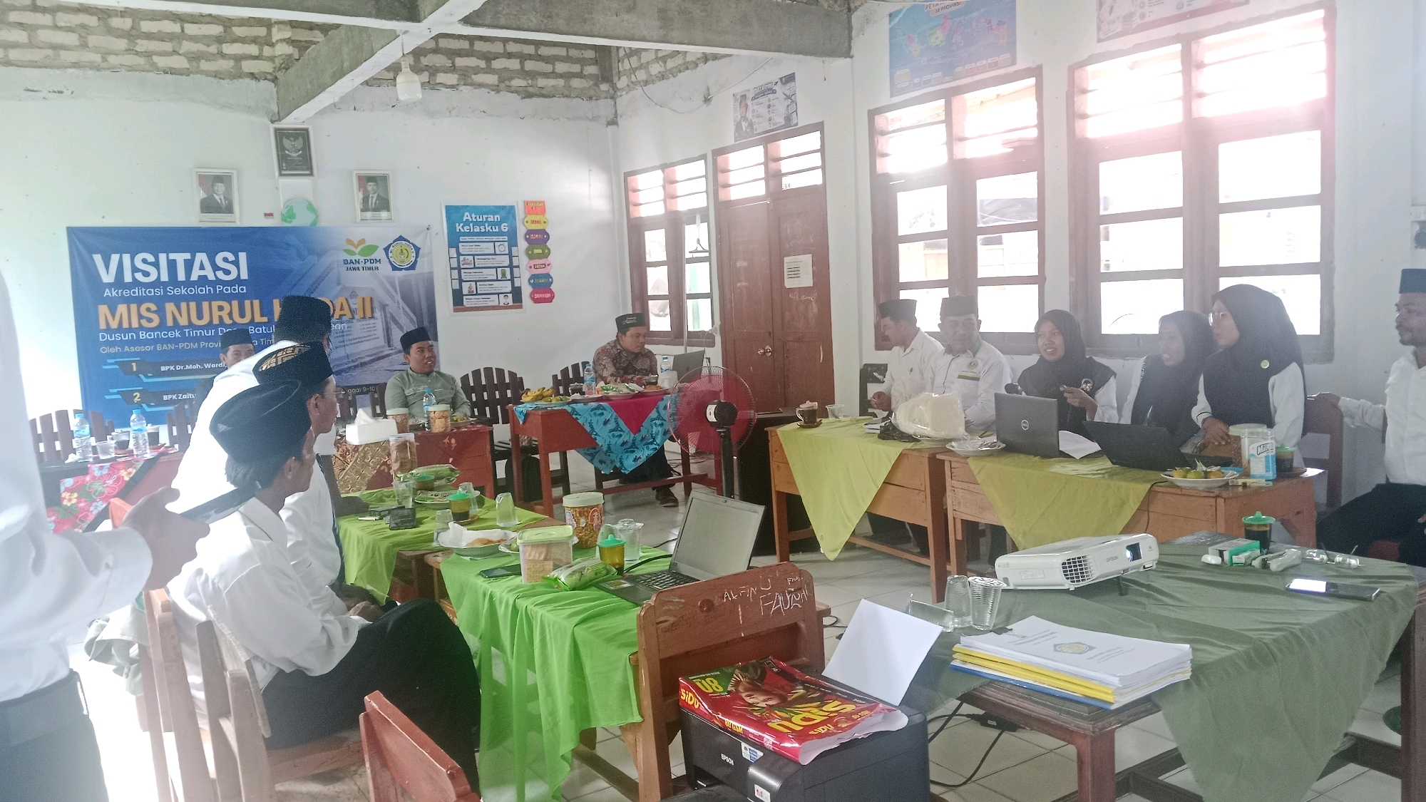 In Dempo Barat, Indonesia, a group of people, likely educators and visitors, are gathered in a room that appears to be a classroom or meeting hall. The room has a rustic feel with a concrete ceiling and large windows that let in natural light. A banner in the background reads "VISITASI AKREDITASI SEKOLAH PADA MIS NURUL HIDAII," indicating an accreditation visit for a school.
Several individuals are seated at tables covered with green cloths. Some are wearing white shirts and caps, common attire in religious or educational settings in Indonesia. Others are wearing hijabs. One person in the foreground is partially visible, wearing a white shirt and looking towards the group.
On the tables, there are various items such as laptops, water bottles, food, and stacks of papers. A projector is set up on one of the tables, suggesting a presentation is taking place or has just concluded. A red electric fan is also visible. The room is decorated with posters on the walls, including one titled "Aturan Kelasku 6" (My Class Rules 6). The overall atmosphere seems to be one of a formal meeting or evaluation.