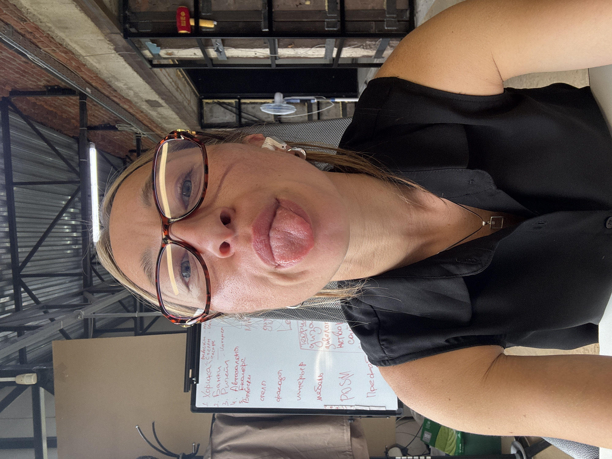 This media file shows a woman with glasses sticking her tongue out at the camera. She is wearing a black shirt and a necklace. She appears to be indoors in a workshop or studio space, with exposed brick and industrial-style metal structures visible in the background. A whiteboard with handwritten text in Russian is partially visible to her right, featuring words like "Вывески" (signs), "Банки" (banks), "интерьер" (interior), and "мебель" (furniture). The lighting suggests it is daytime. The woman's playful expression indicates a casual and lighthearted mood. The text on the whiteboard might be related to a business or project, possibly for interior design or marketing given the terms present.