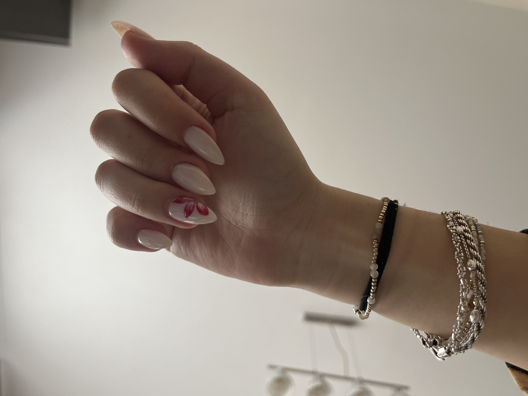 A woman's hand is shown from a low angle, with the hand pointed upward. The hand is fair-skinned with almond-shaped nails, which are light in color. One nail has a small red flower design. On the wrist, there are three bracelets: one with gold beads and a black band, and two layered silver bracelets. The background is a plain, light-colored wall and a blurry image of a modern light fixture. No specific information is available to definitively place the scene in Sofia, Bulgaria.
