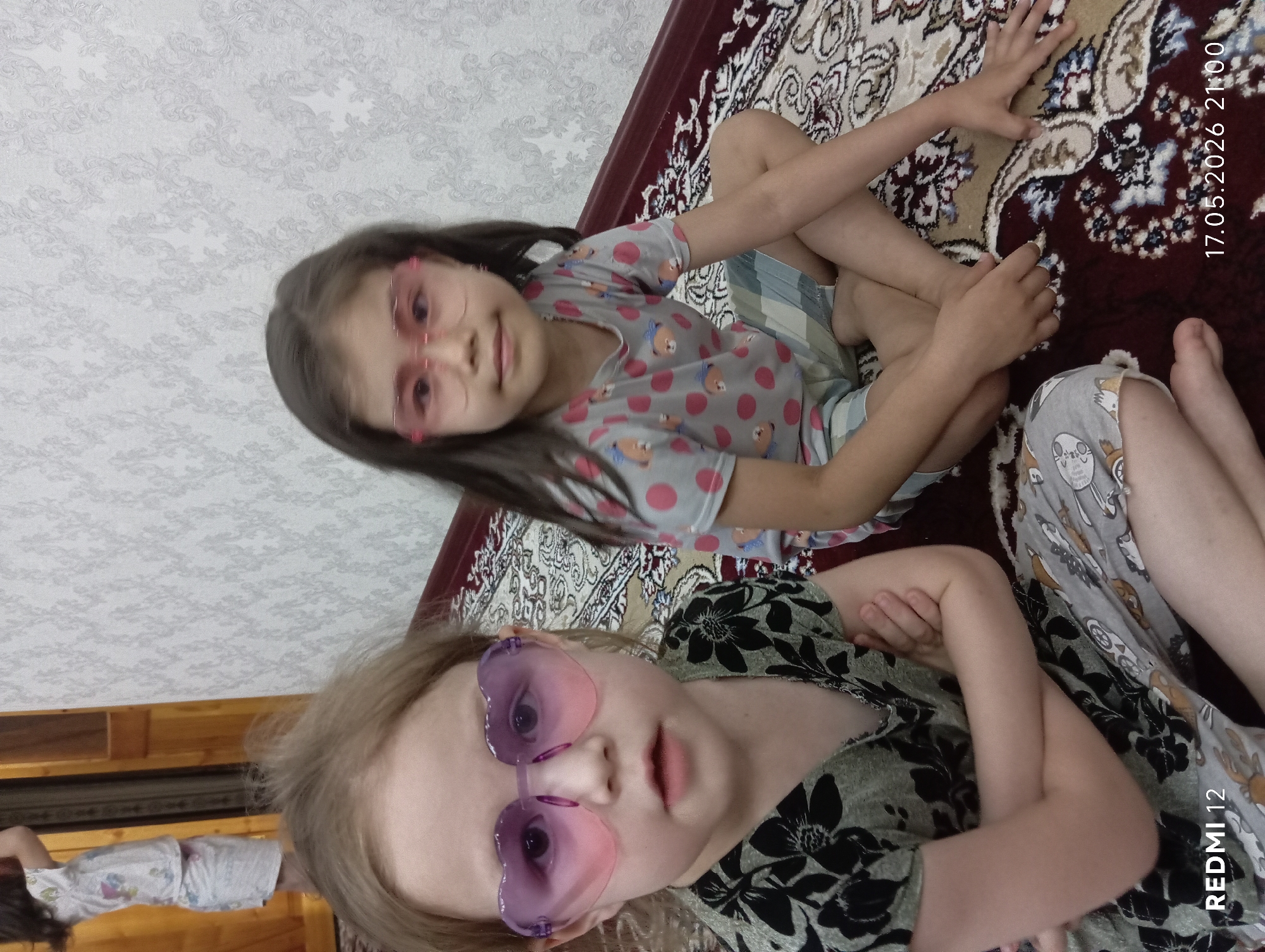 Two young girls are sitting on a patterned rug. The girl in the foreground is wearing heart-shaped sunglasses and a floral print top. The other girl is wearing a shirt with teddy bear patterns and is wearing light-colored framed glasses. The date "17.05.2026 21:00" is visible in the upper right corner. The scene is in Bekabad, Uzbekistan.