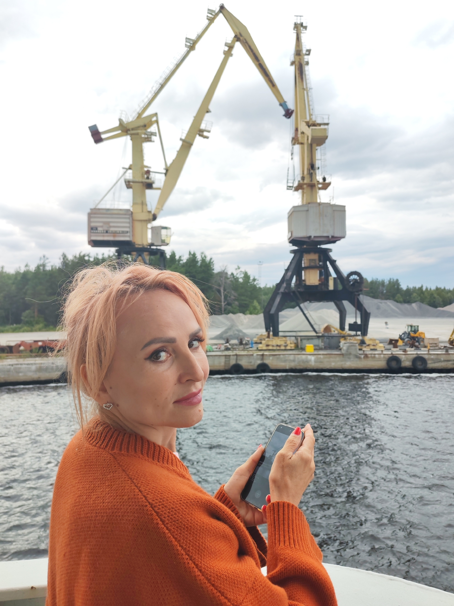 A woman with peach-colored hair, wearing an orange sweater, is holding a smartphone in her hands. She is looking over her shoulder at the camera. In the background, there are two large yellow cranes, a body of water, and a cloudy sky. The scene appears to be a port or industrial area, possibly in Shalsky, Russia, given the presence of heavy machinery like cranes and what looks like piles of gravel or aggregate. The woman is positioned in the foreground, with the industrial landscape and cranes occupying the midground and background.