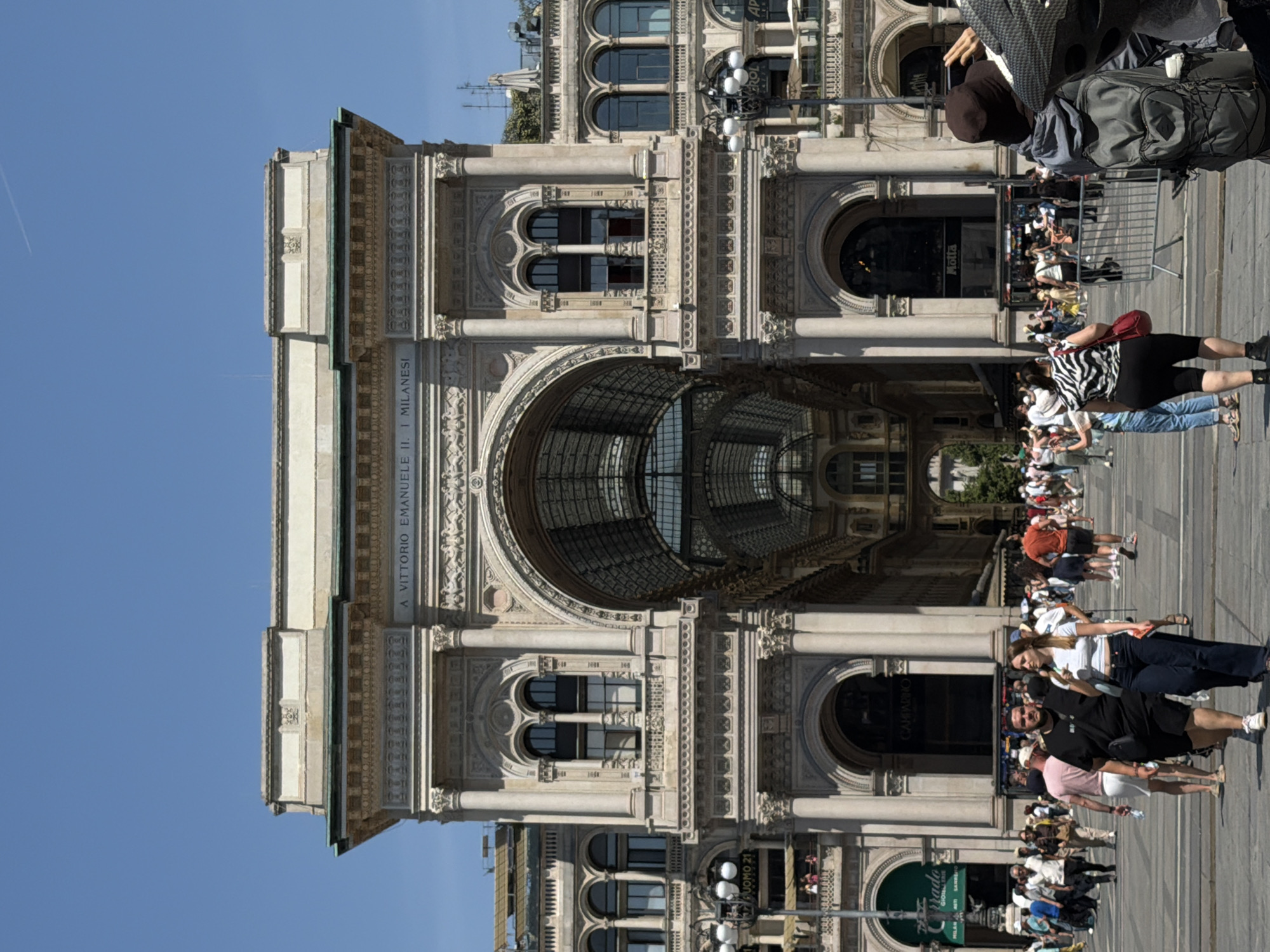 This image captures the Galleria Vittorio Emanuele II, a famous shopping arcade in Milan, Italy. The arcade's architecture features a glass-covered vaulted ceiling and ornate stucco decorations. A multitude of people are walking around, some in groups and others individually. They are engaged in activities like shopping, sightseeing, and taking pictures. Sunlight bathes the scene, creating shadows and reflections.